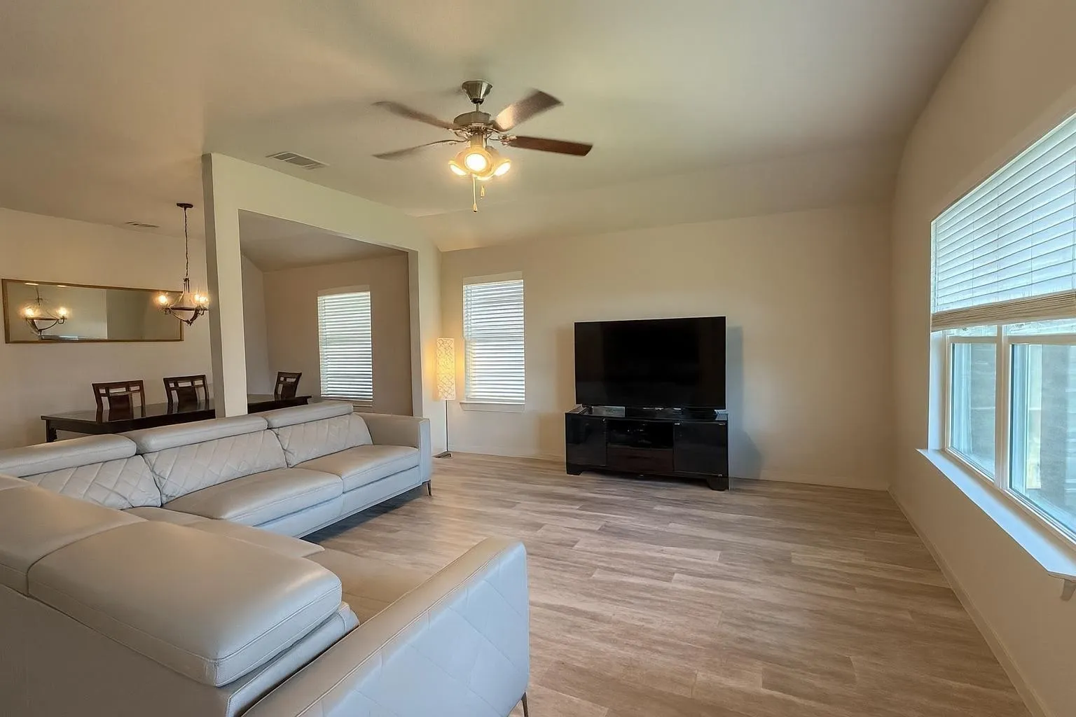 Spacious living area featuring wood-finish flooring, a ceiling fan with integrated lighting, and multiple windows with blinds