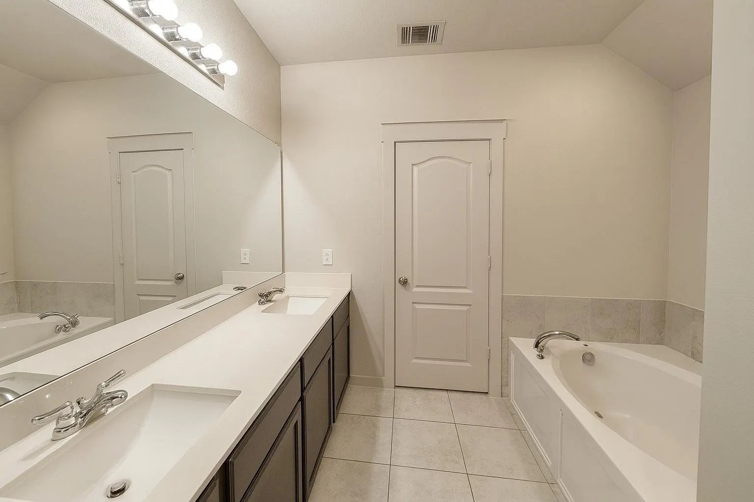 Bathroom featuring a dual vanity with undermount sinks, a large integrated mirror, overhead lighting, and a built-in bathtub with a tiled surround