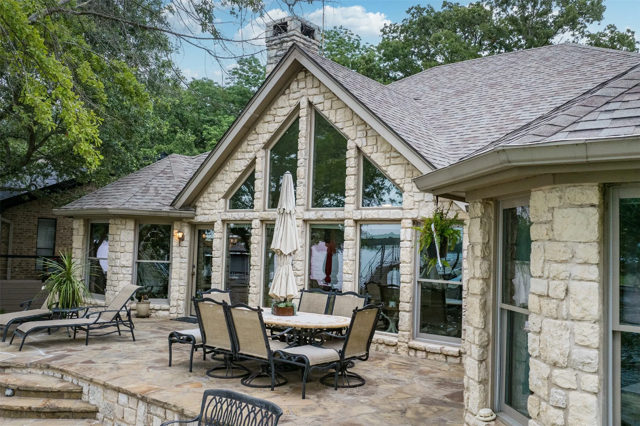 Stone exterior with a prominent gable, expansive windows, a multi-level stone patio, and a shingle roof