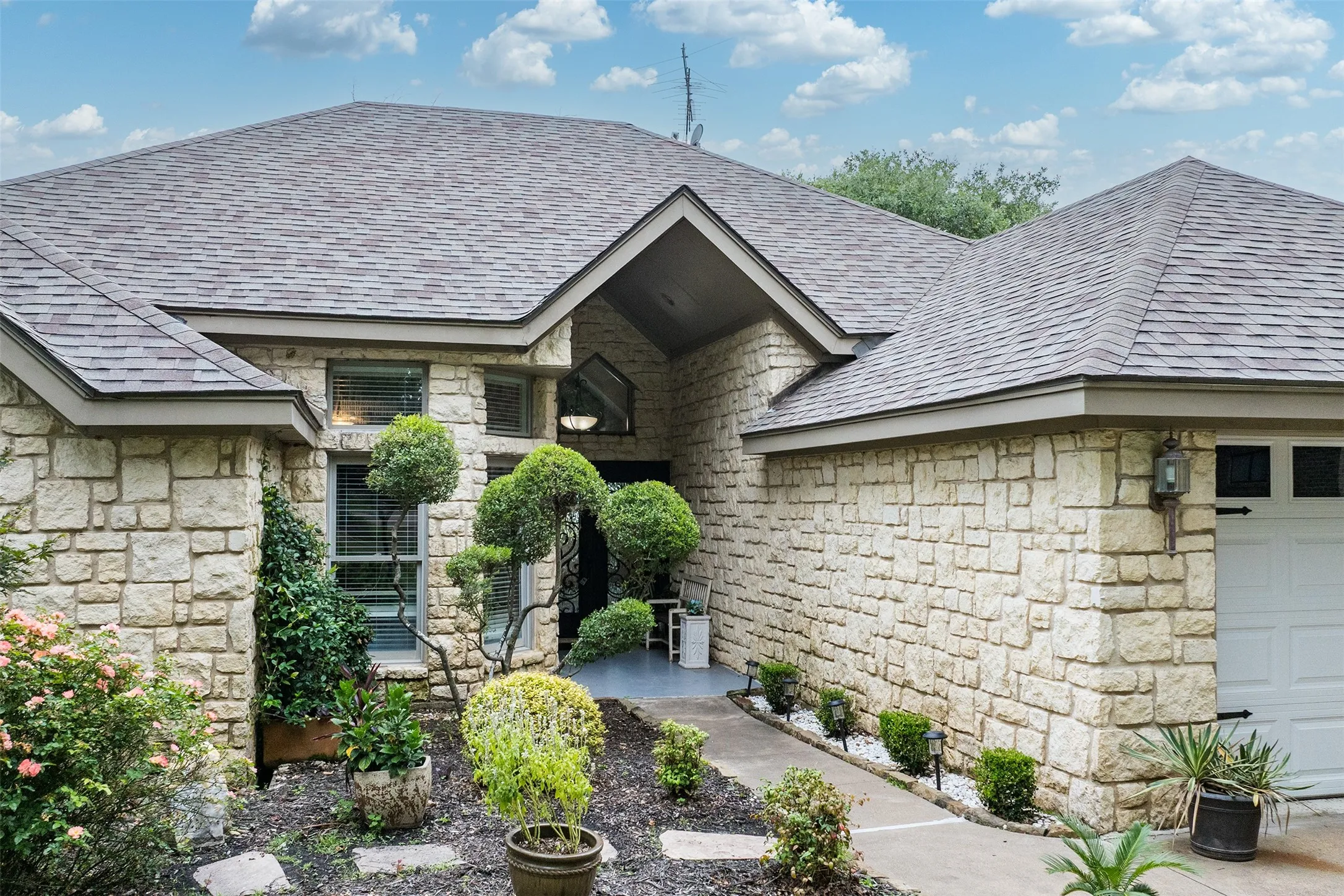 Stone façade residence featuring a multi-gabled shingle roof, a recessed entry with an arched transom window, a concrete pathway, and a two-car garage with overhead lighting