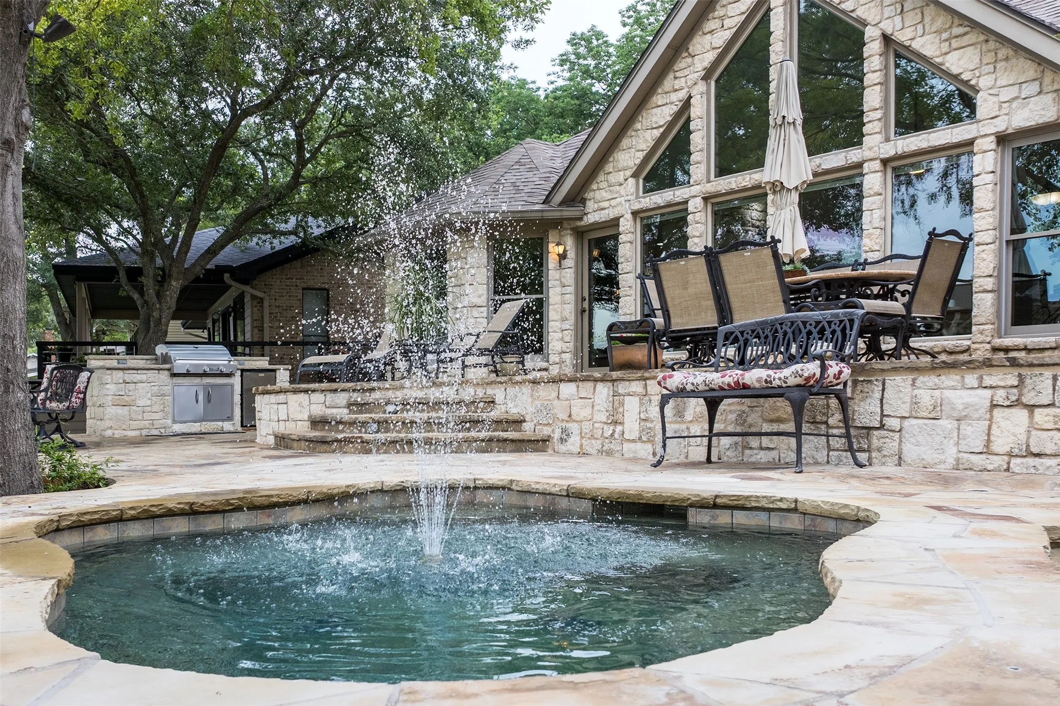 Stone exterior showcasing an outdoor water feature with a fountain, a built-in outdoor grill station, a covered patio, and large, angular windows