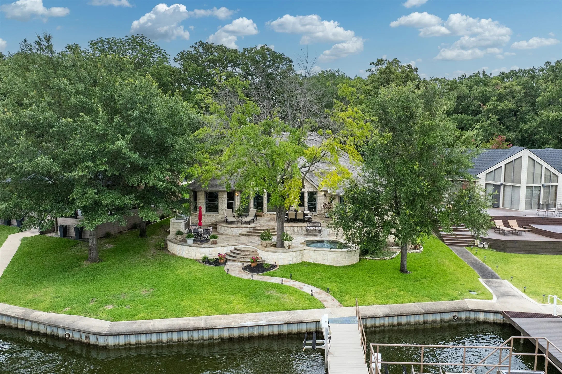 Waterfront property featuring a private dock, tiered patio, built-in hot tub, and lush green lawn