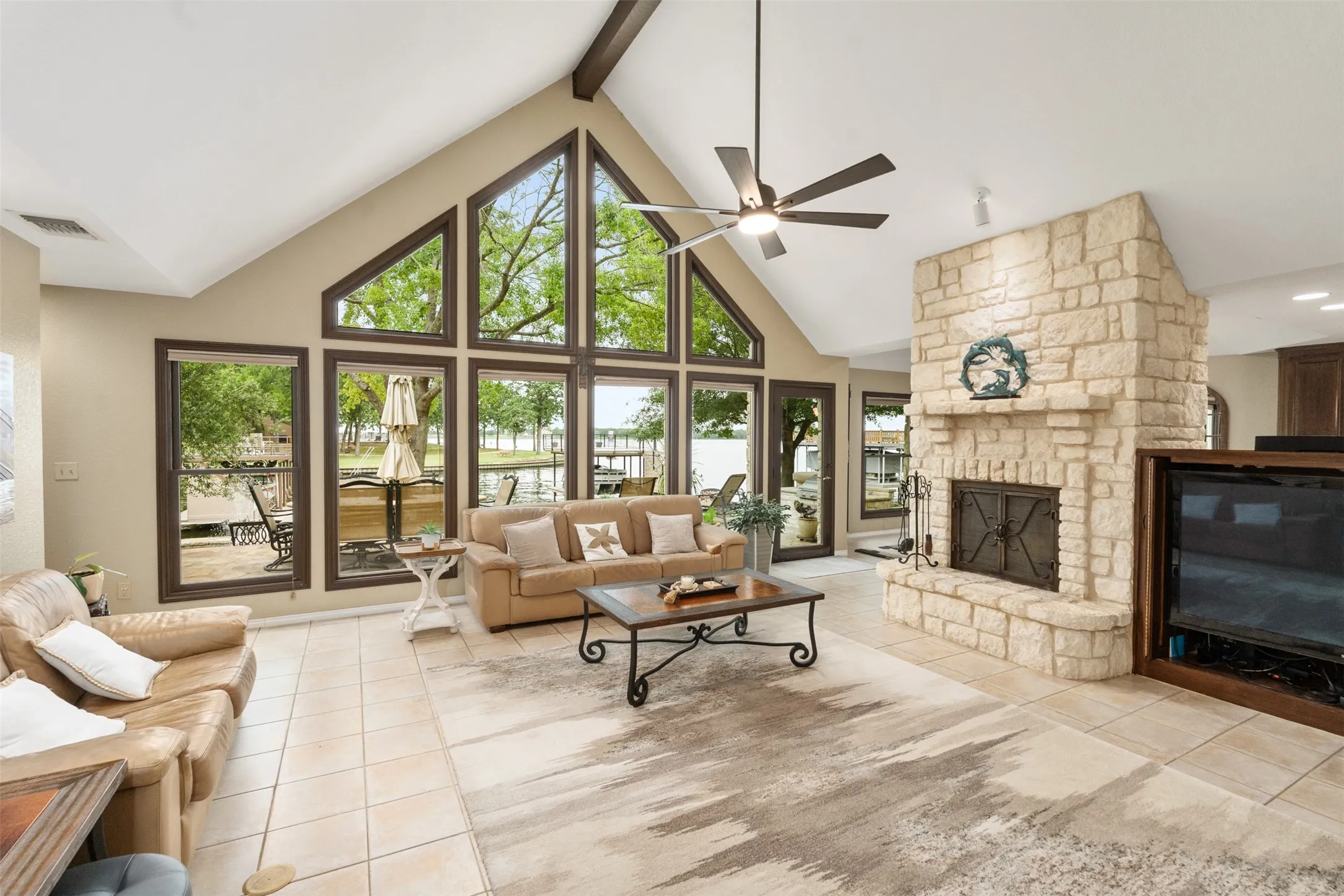 Vaulted ceiling living area featuring a stone fireplace, ceramic tile flooring, ceiling fan, and extensive picture windows offering waterfront views