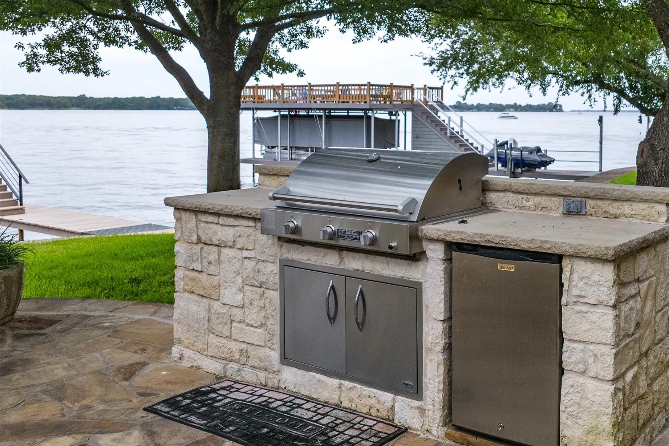 Outdoor kitchen featuring a built-in stainless steel grill, integrated mini-refrigerator, and stone-clad base with a solid countertop