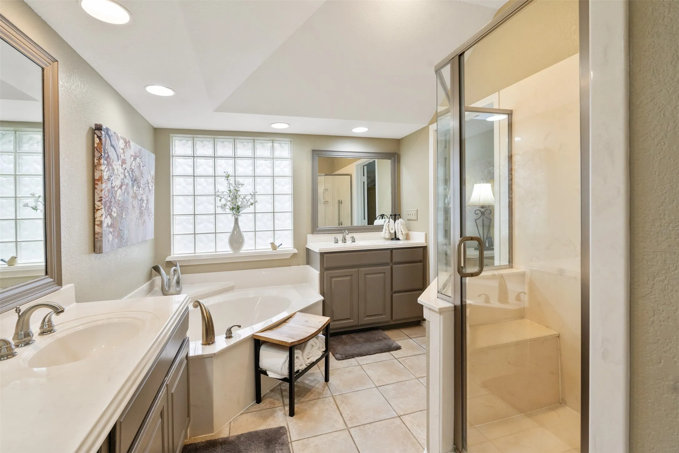 Spacious bathroom featuring a built-in corner soaking tub, a glass-enclosed shower with a built-in bench, dual vanity sinks with light-toned countertops, and recessed lighting