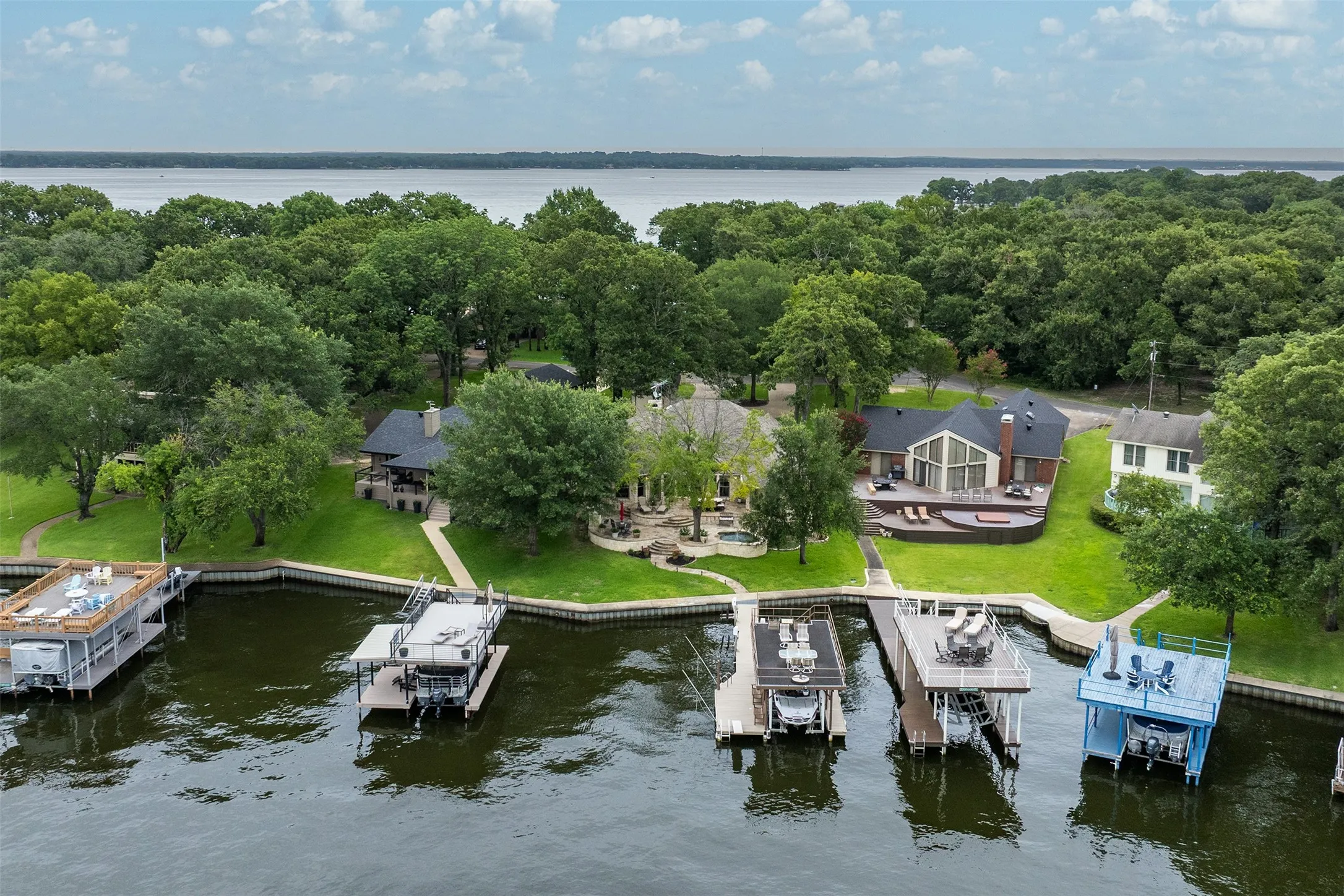 Waterfront property featuring multiple private boat docks with upper decks, a manicured lawn, mature trees, and a spacious rear deck with a built-in hot tub