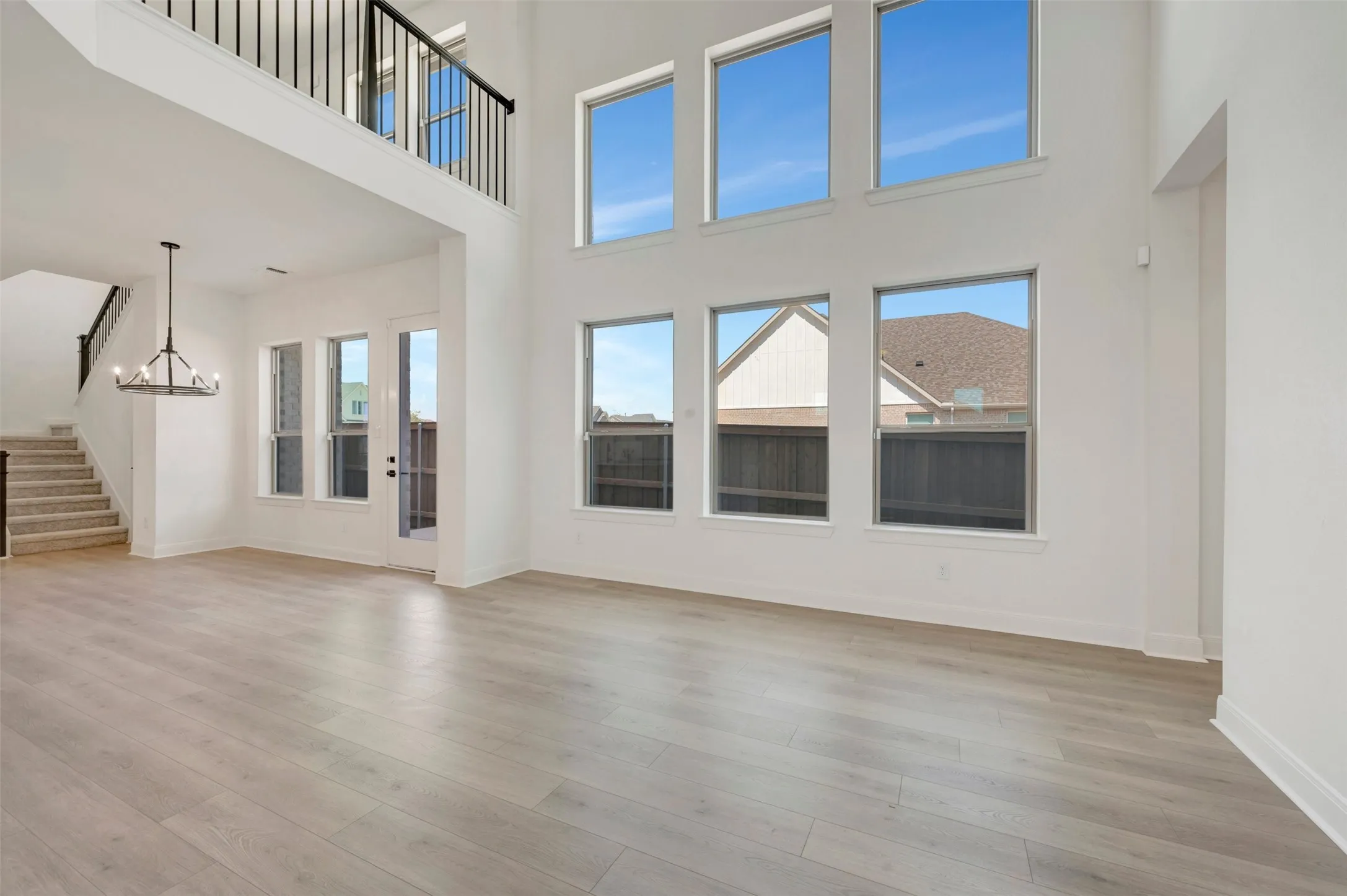Two-story living area with high ceilings, wood-finish flooring, a contemporary chandelier, multiple windows, and a second-story balcony with metal railings