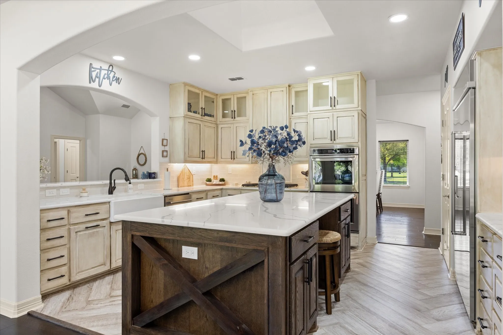 Bright kitchen featuring a central island with a white countertop, light-toned cabinetry, stainless steel oven, and a farmhouse sink