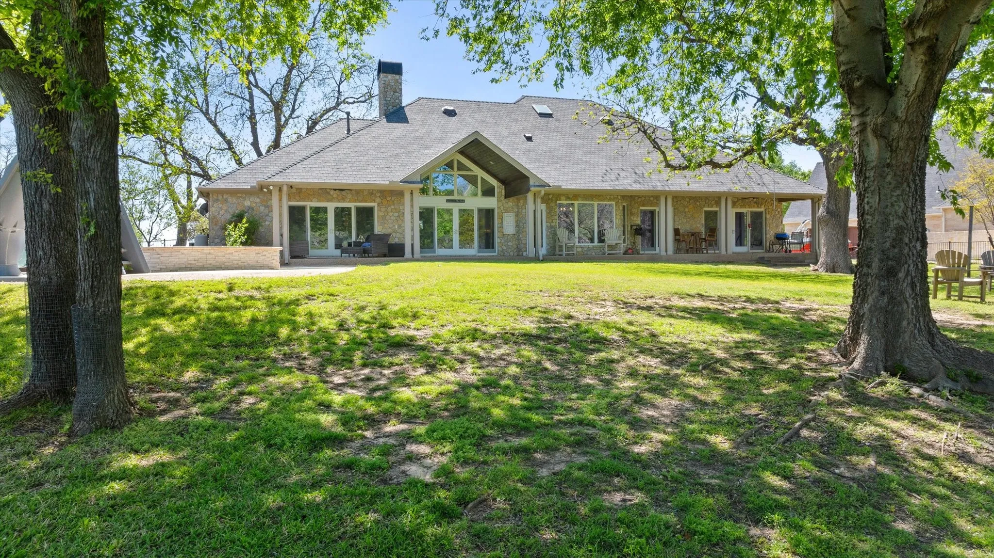 Expansive rear exterior featuring a stone facade, multiple sets of glass doors, a covered patio extending the length of the home, and a pitched roofline with a prominent chimney