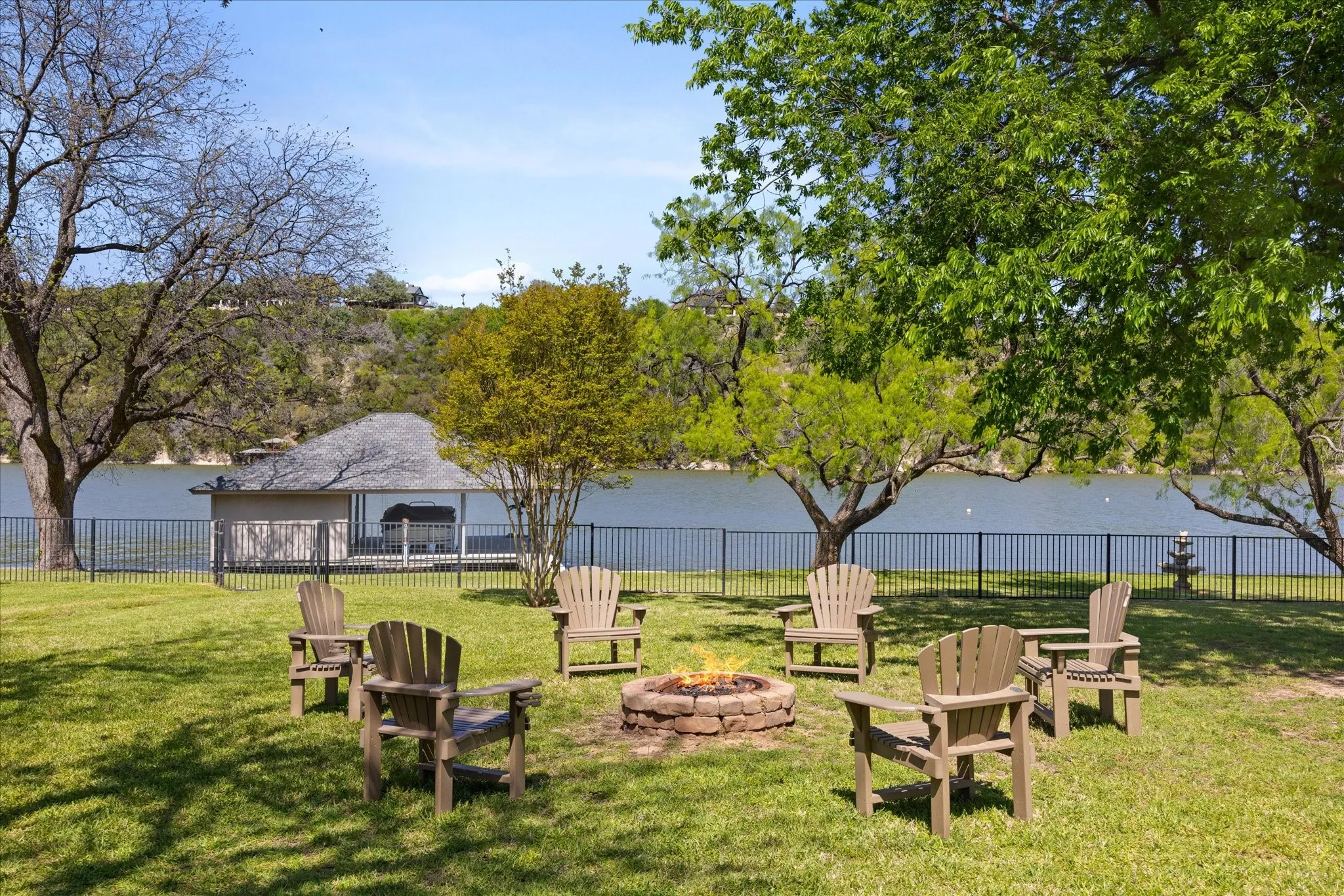 Lush green lawn with a built-in stone fire pit, mature trees, and a black wrought iron fence
