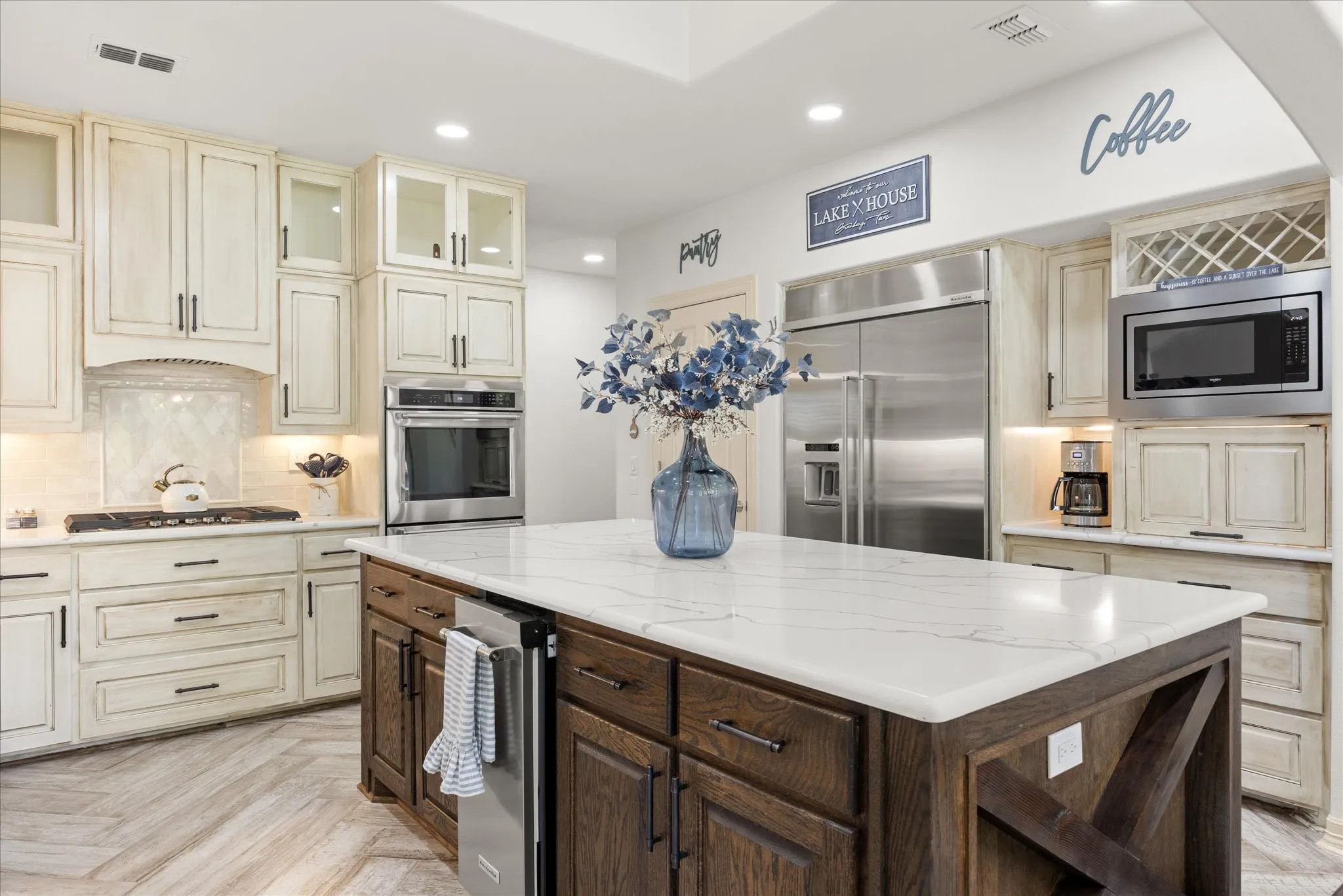 Kitchen featuring a large central island with a light-toned countertop, stainless steel appliances, distressed white cabinetry, wood-finish flooring, and recessed lighting