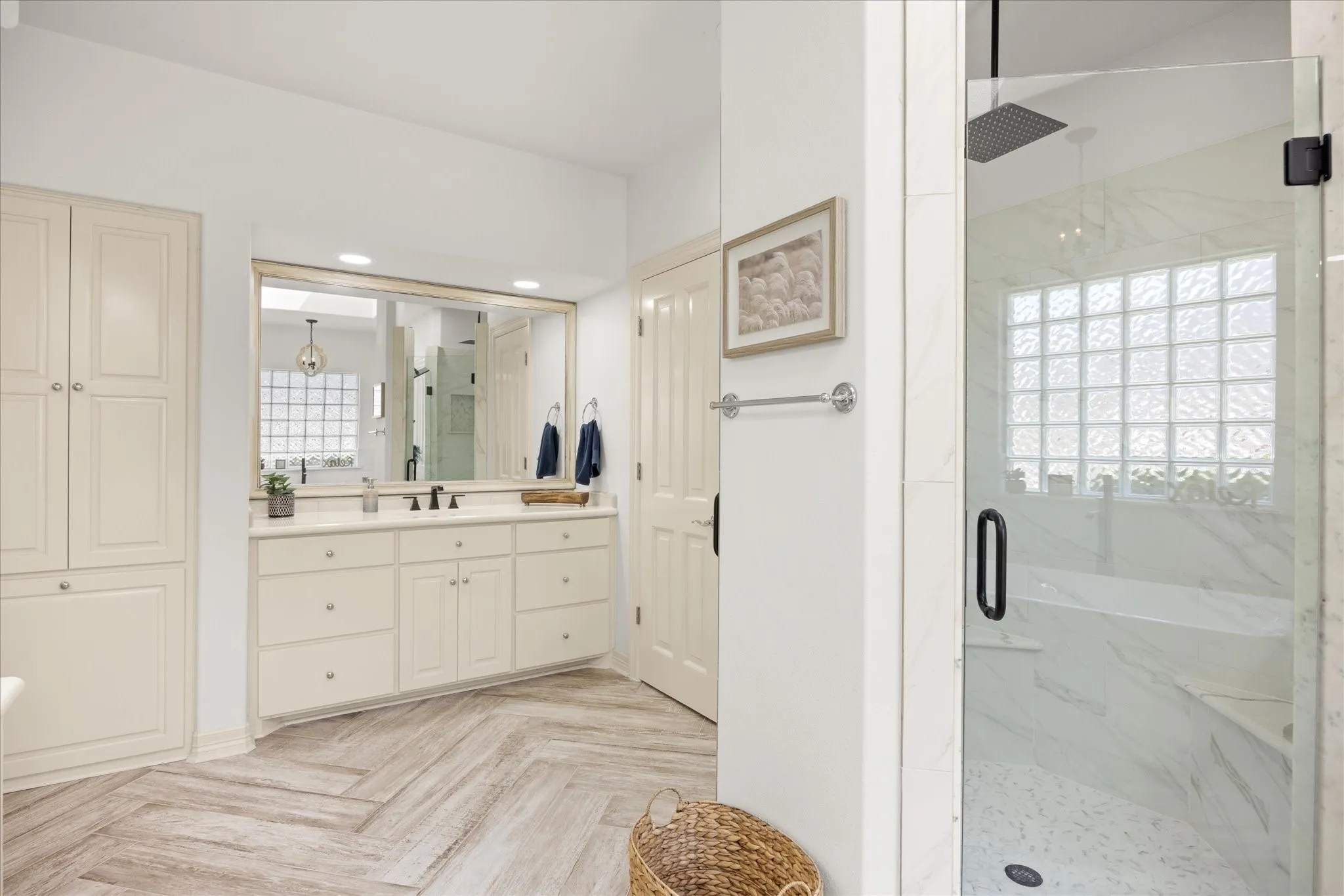 Spacious bathroom featuring a glass-enclosed shower with a bench, a double vanity with a quartz countertop, cream-colored cabinetry, built-in storage, and wood-finish flooring laid in a herringbone pattern