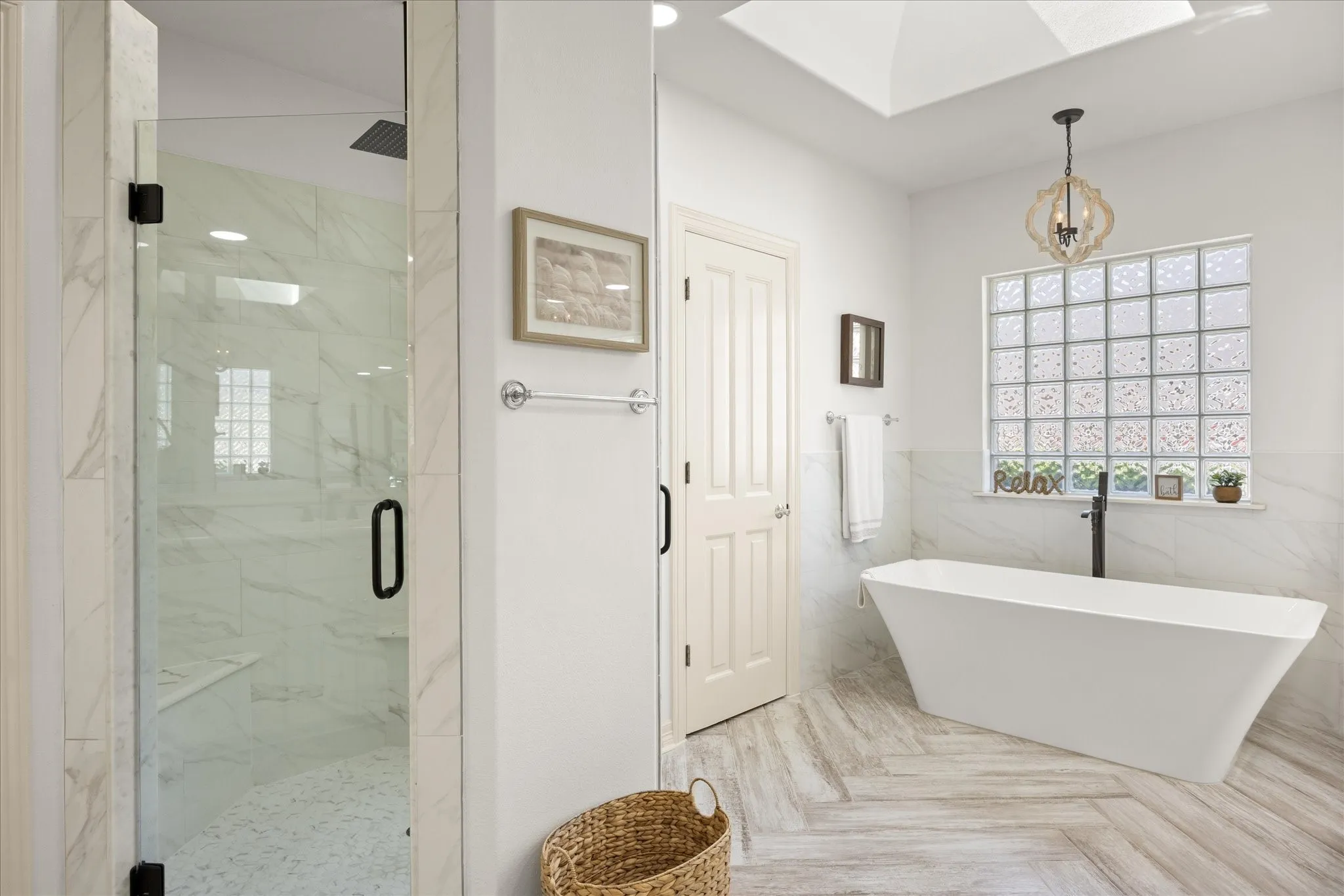 White freestanding soaking tub with a floor-mounted black faucet, glass block window, wood-finish herringbone flooring, and a glass-enclosed shower with a built-in bench