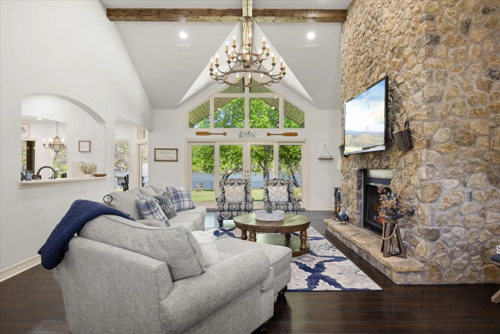 Vaulted ceilings with exposed wood beams, a stone-clad fireplace with a hearth, full-height windows, a prominent chandelier, and dark wood-finish flooring