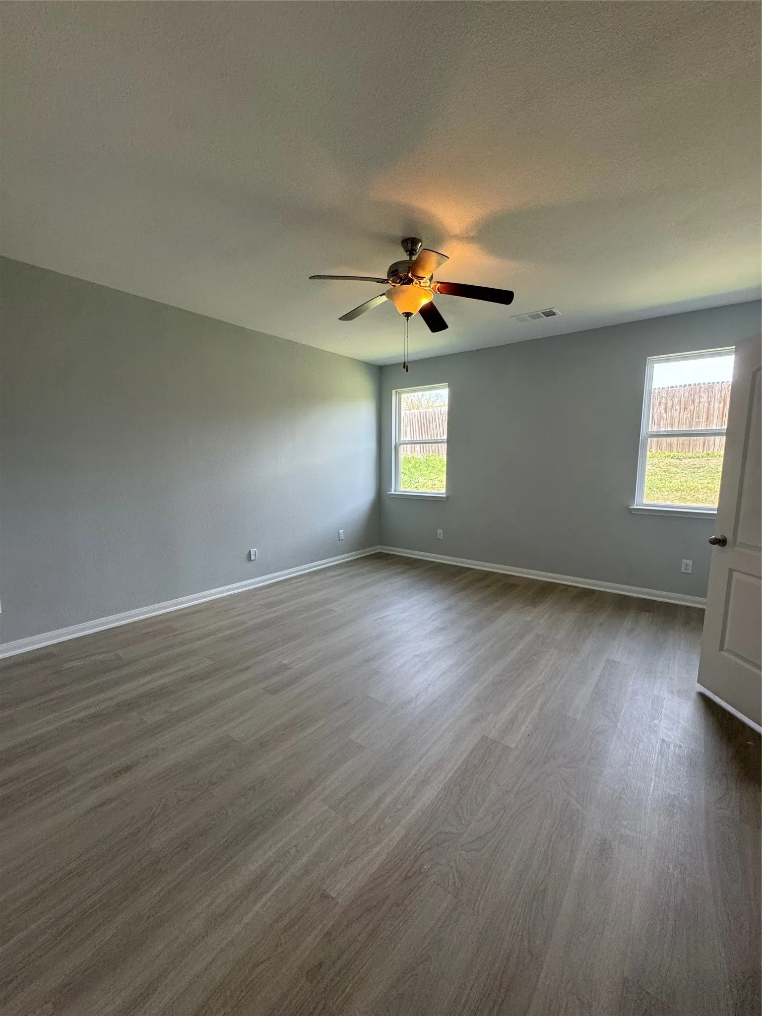 Spacious room featuring wood-finish flooring, two windows, a ceiling fan with integrated lighting, and neutral wall tones