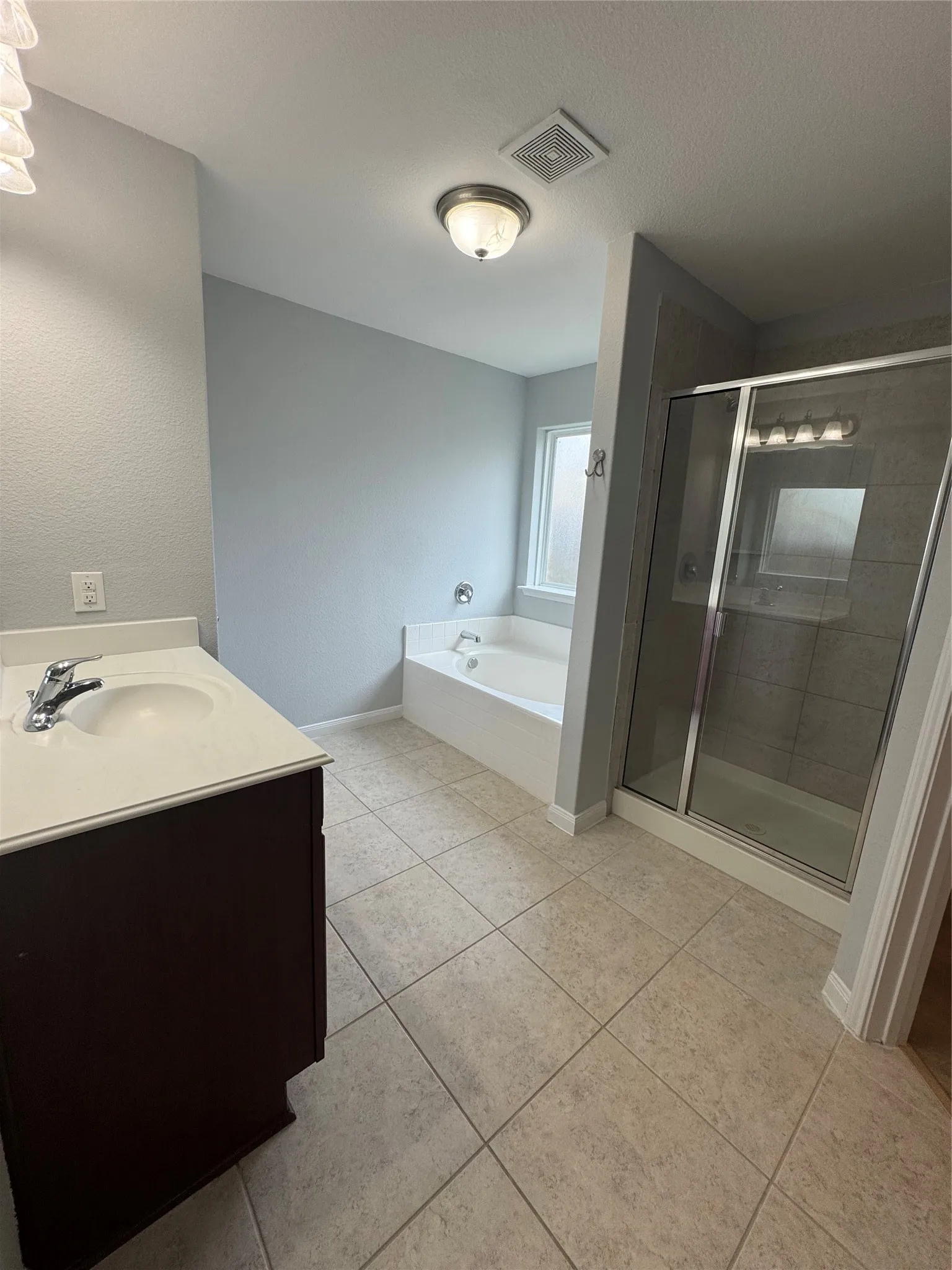 Bathroom featuring a single vanity with a white countertop and dark wood-finish cabinetry, a built-in corner soaking tub, a glass-enclosed shower with tiled walls, and light-toned tile flooring
