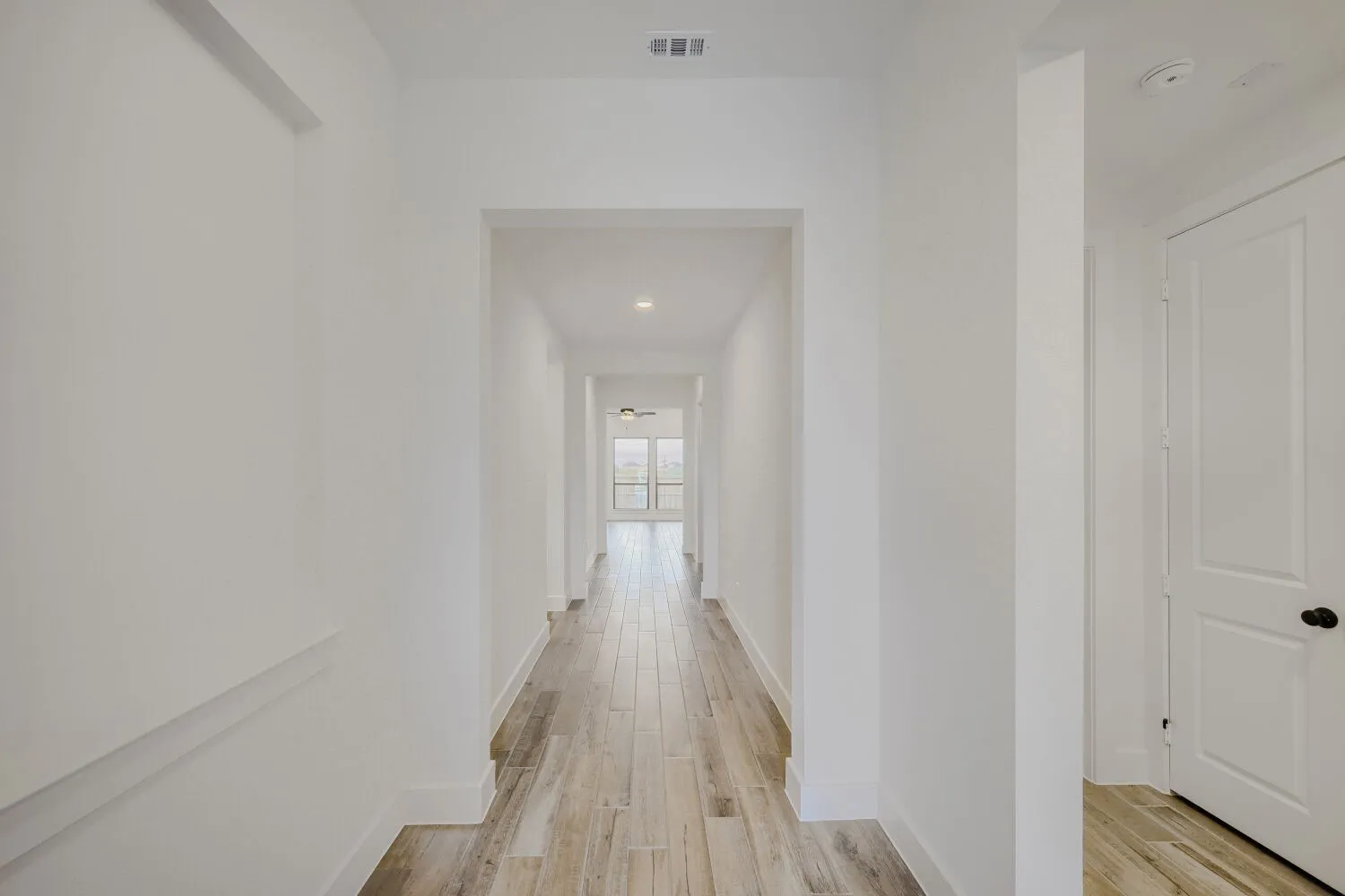 Hallway with wood-finish flooring extending into the background, bright white walls, recessed ceiling lighting, and a paneled door with dark hardware