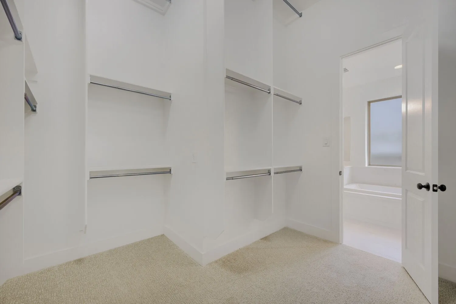 Spacious closet featuring multiple shelving units with hanging rods, light-toned carpeting, white baseboards, and a doorway leading to a bathroom with a window and a built-in tub