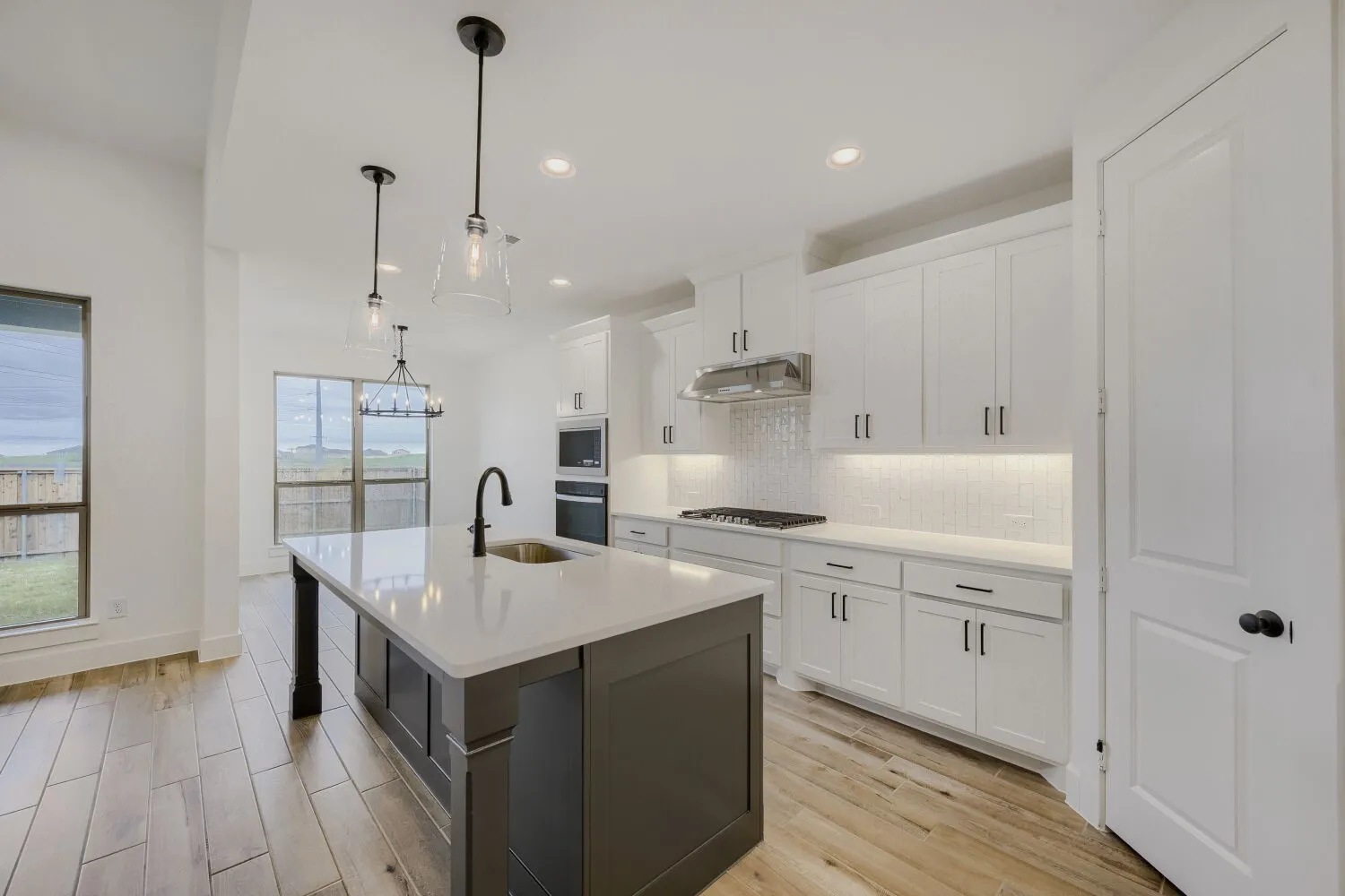 Kitchen featuring a central island with a sink and modern faucet, light-toned wood-finish flooring, white cabinetry with dark hardware, built-in stainless steel appliances, and pendant lighting