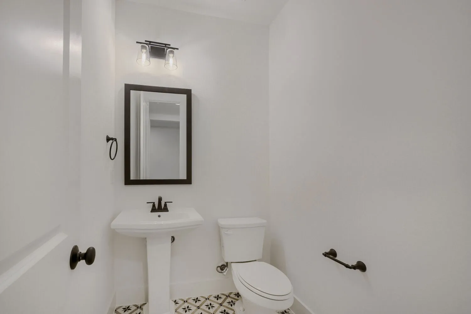 Pedestal sink with a black-framed rectangular mirror, black wall-mounted light fixture, and patterned floor tile
