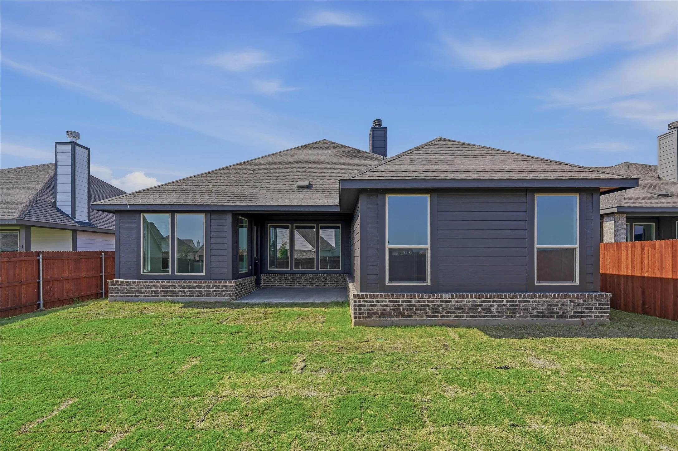 Expansive backyard featuring a manicured lawn, dark siding, brick detailing, and a concrete patio