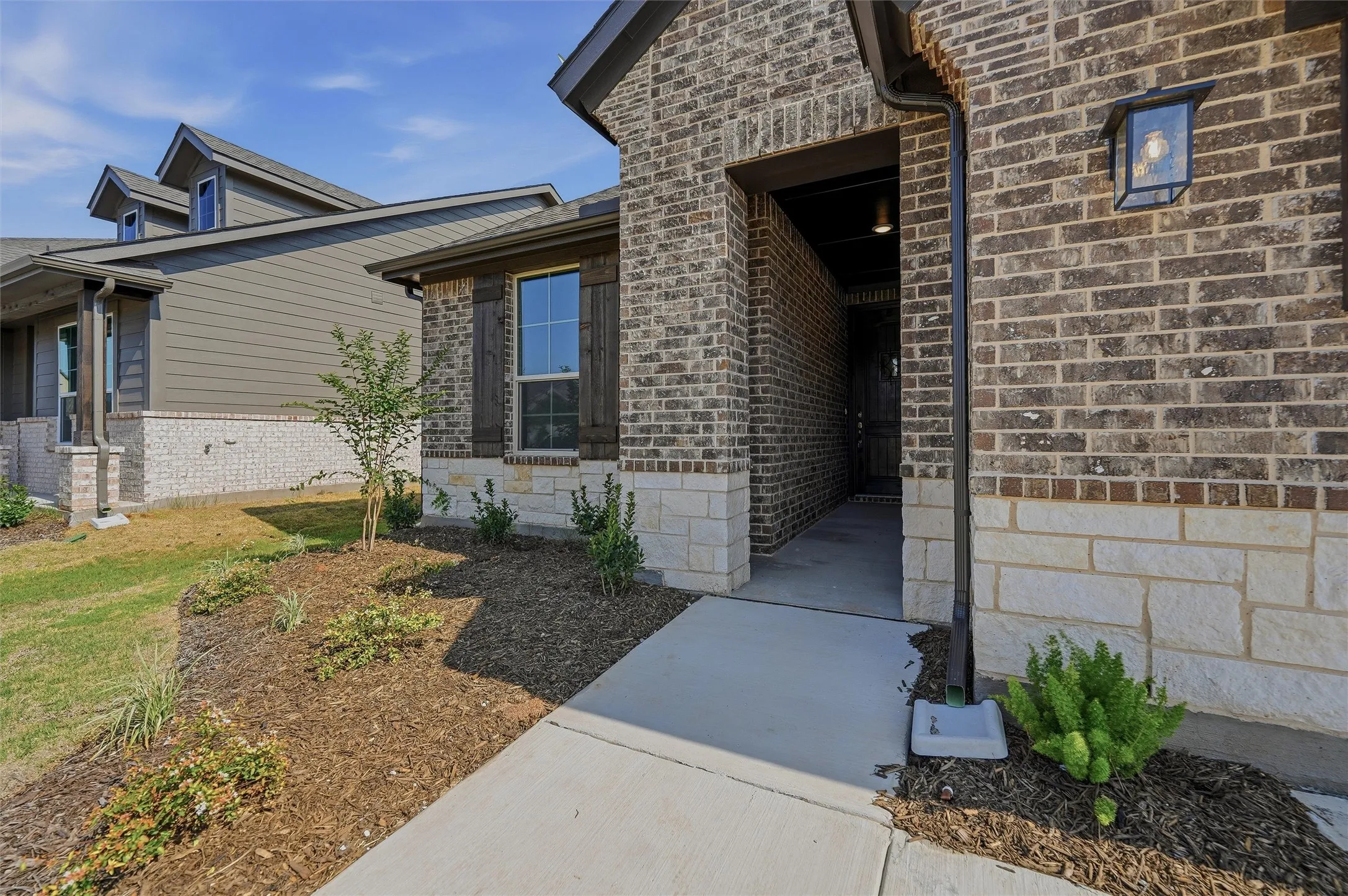 Brick and stone facade, covered entry, decorative exterior lighting, front yard landscaping, and dark-stained window shutters