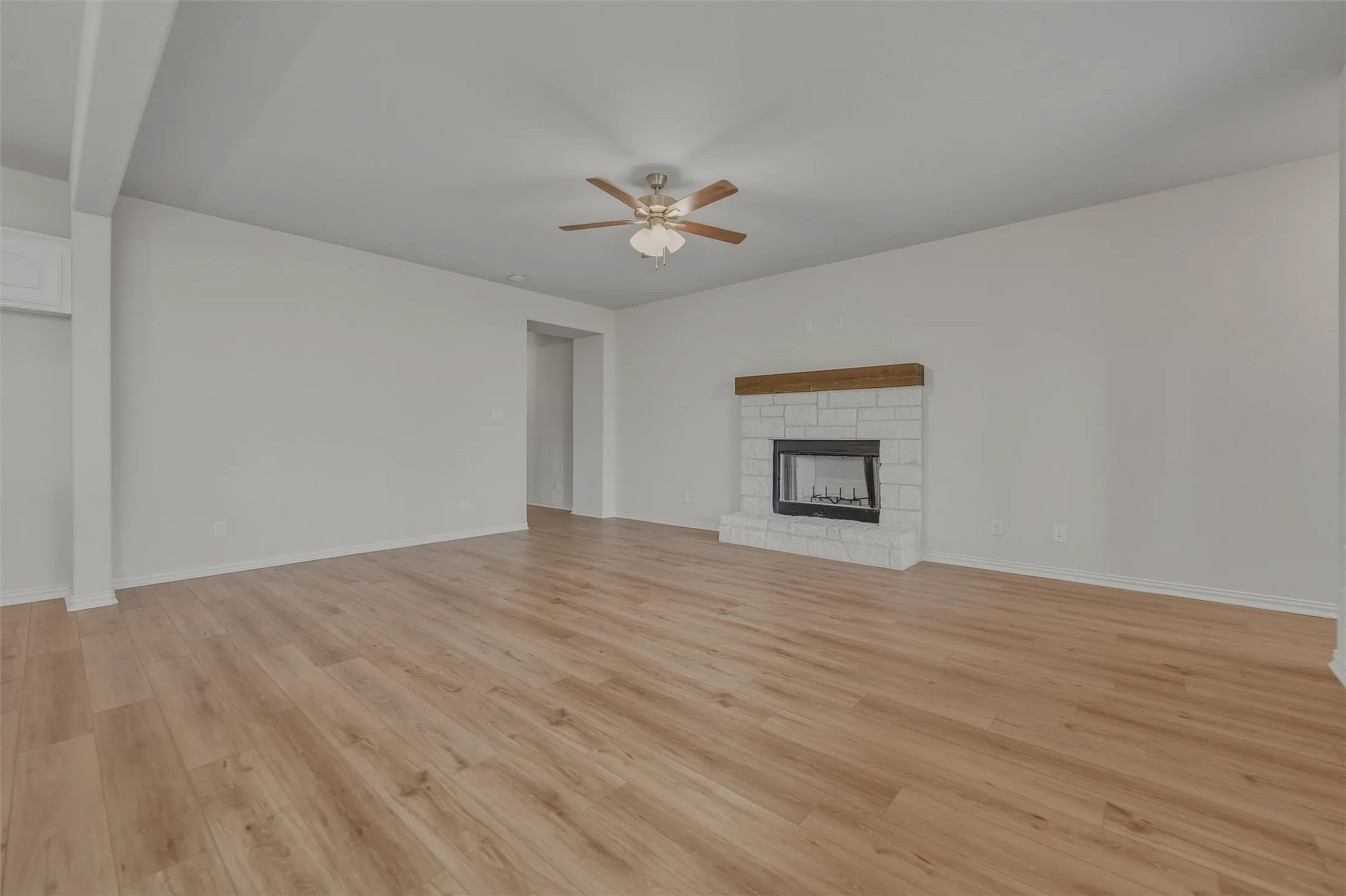 Spacious room featuring wood-finish flooring, a white brick fireplace with a wood mantel, a ceiling fan, and white walls