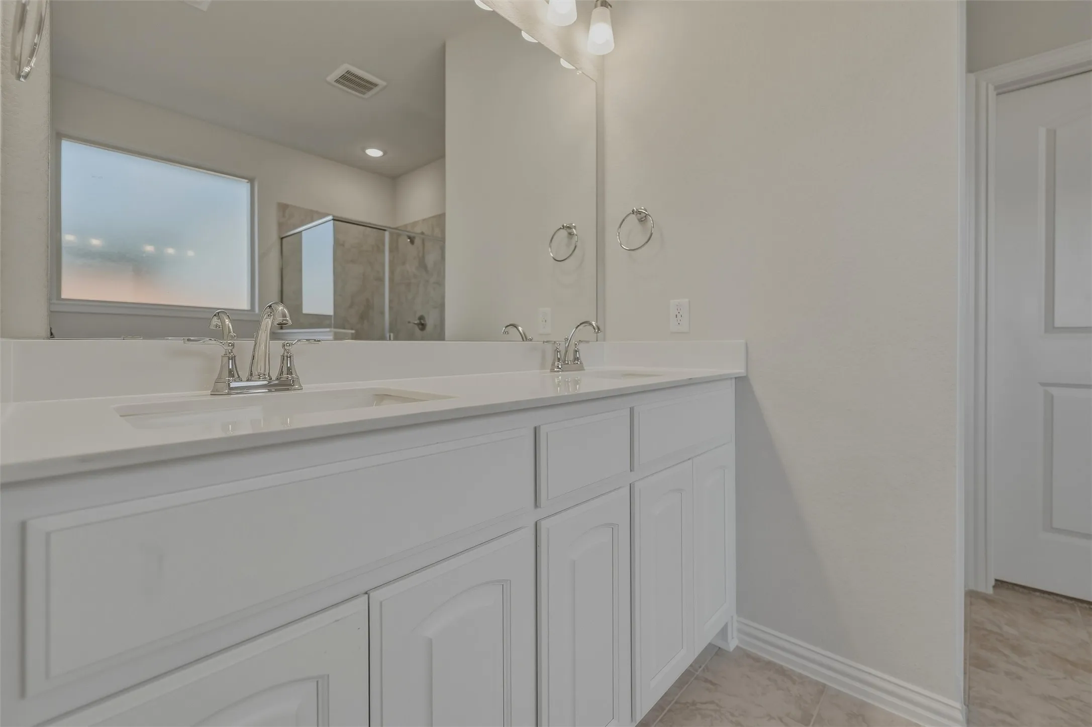 Spa-inspired primary bathroom featuring a white dual-vanity with raised-panel cabinetry, quartz countertops, chrome faucets, and an expansive mirror with integrated lighting