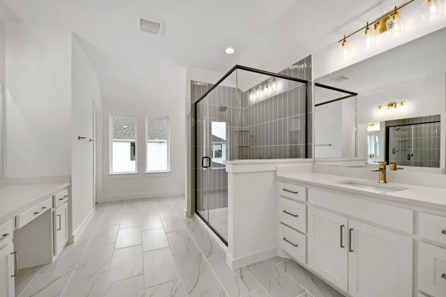 Spacious bathroom featuring a glass-enclosed shower with vertical tile, dual vanities with white cabinetry, gold-finish hardware, and tile flooring