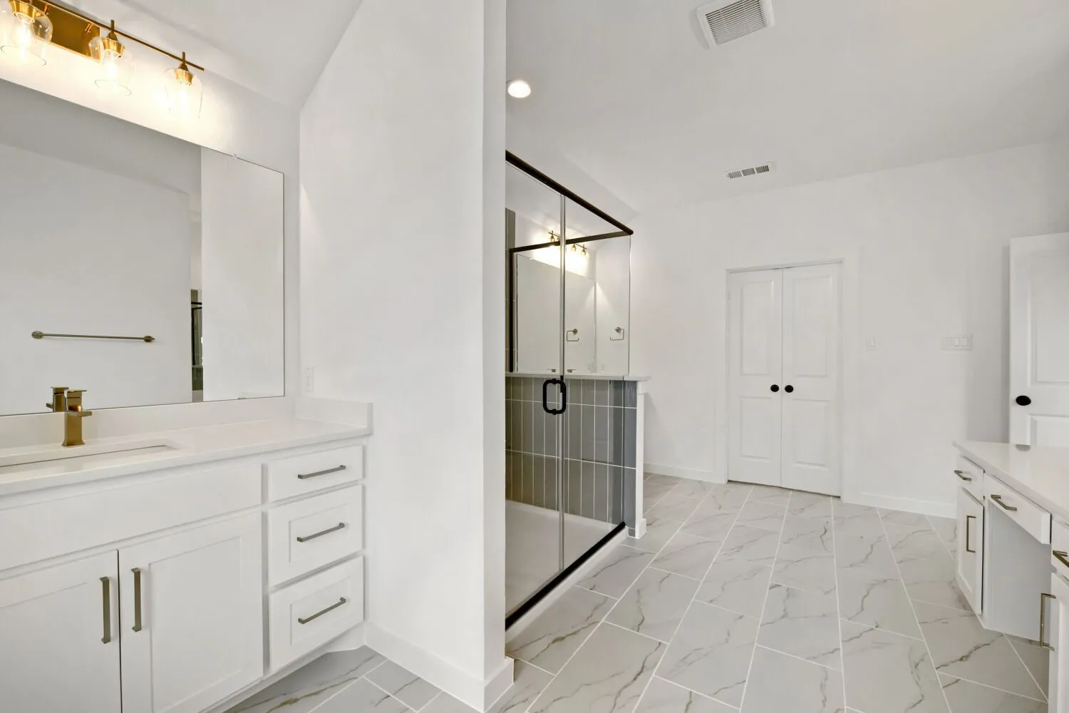 Spacious bathroom featuring a frameless glass shower enclosure, a dual vanity with white cabinetry and light countertops, modern gold-tone fixtures, and large format floor tiles
