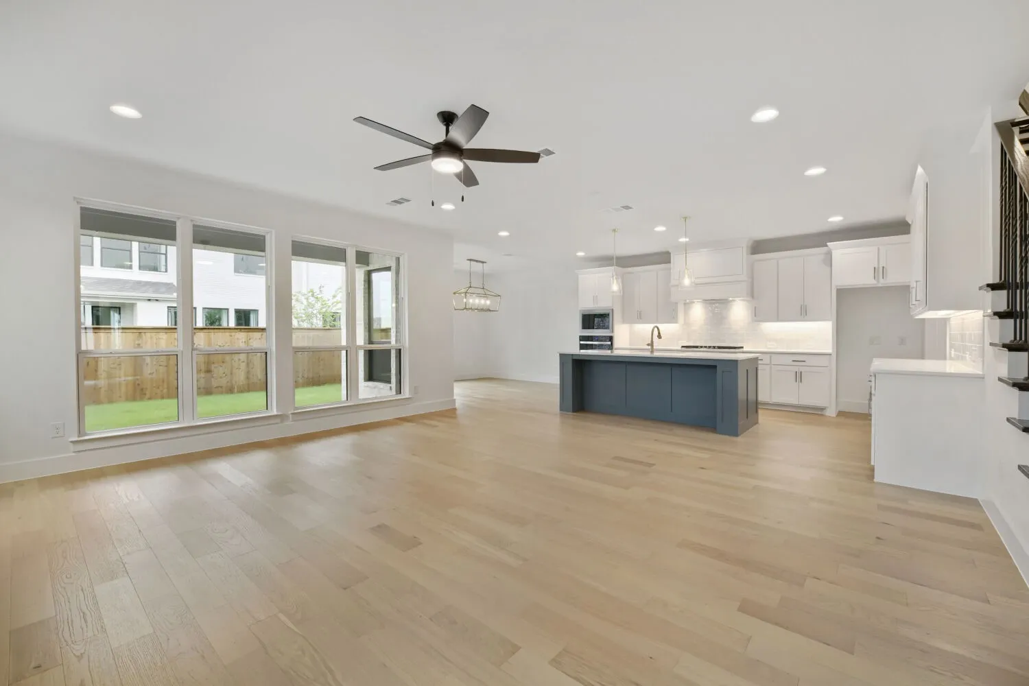 Open-concept living area featuring light wood-finish flooring, a contemporary ceiling fan with integrated lighting, and multiple large windows overlooking a private fenced yard