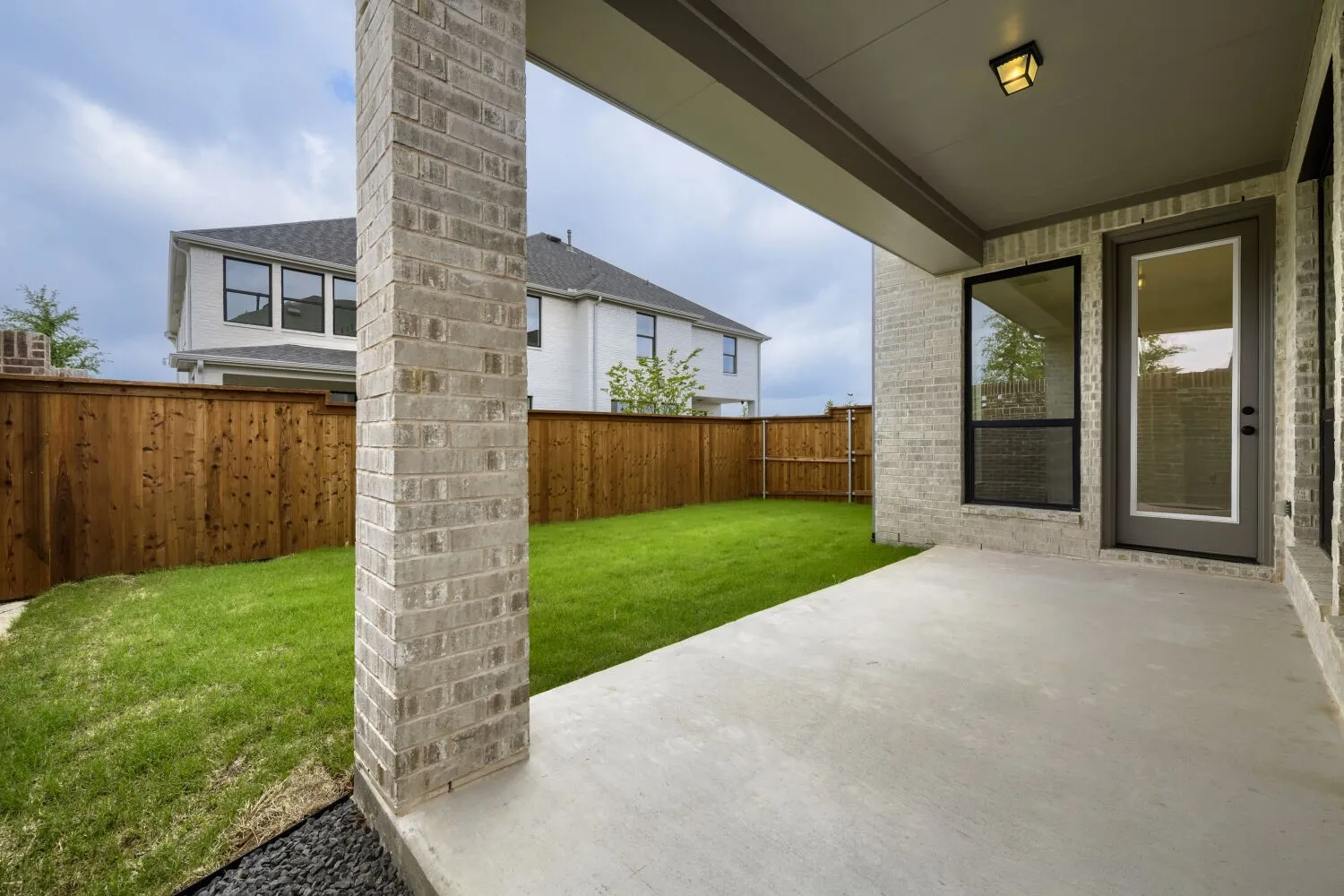 Covered concrete patio with a brick column and exterior wall, featuring a glass-paned door and large window