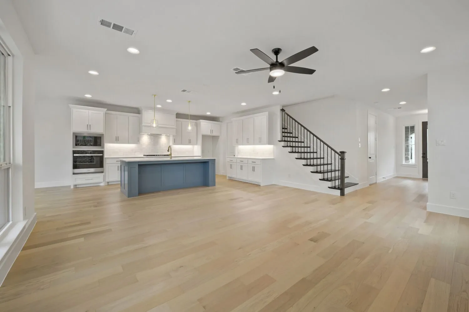 Open-concept living space featuring light wood-finish flooring, a contemporary kitchen with a blue-hued island, white cabinetry, integrated stainless steel appliances, and a staircase with wrought iron balusters