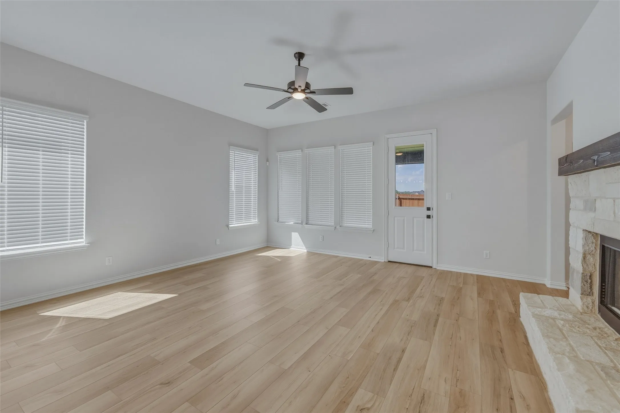 Spacious living area featuring light wood-finish flooring, a stone-clad fireplace with a wood mantle, multiple windows with blinds, a ceiling fan with integrated lighting, and a rear exit door