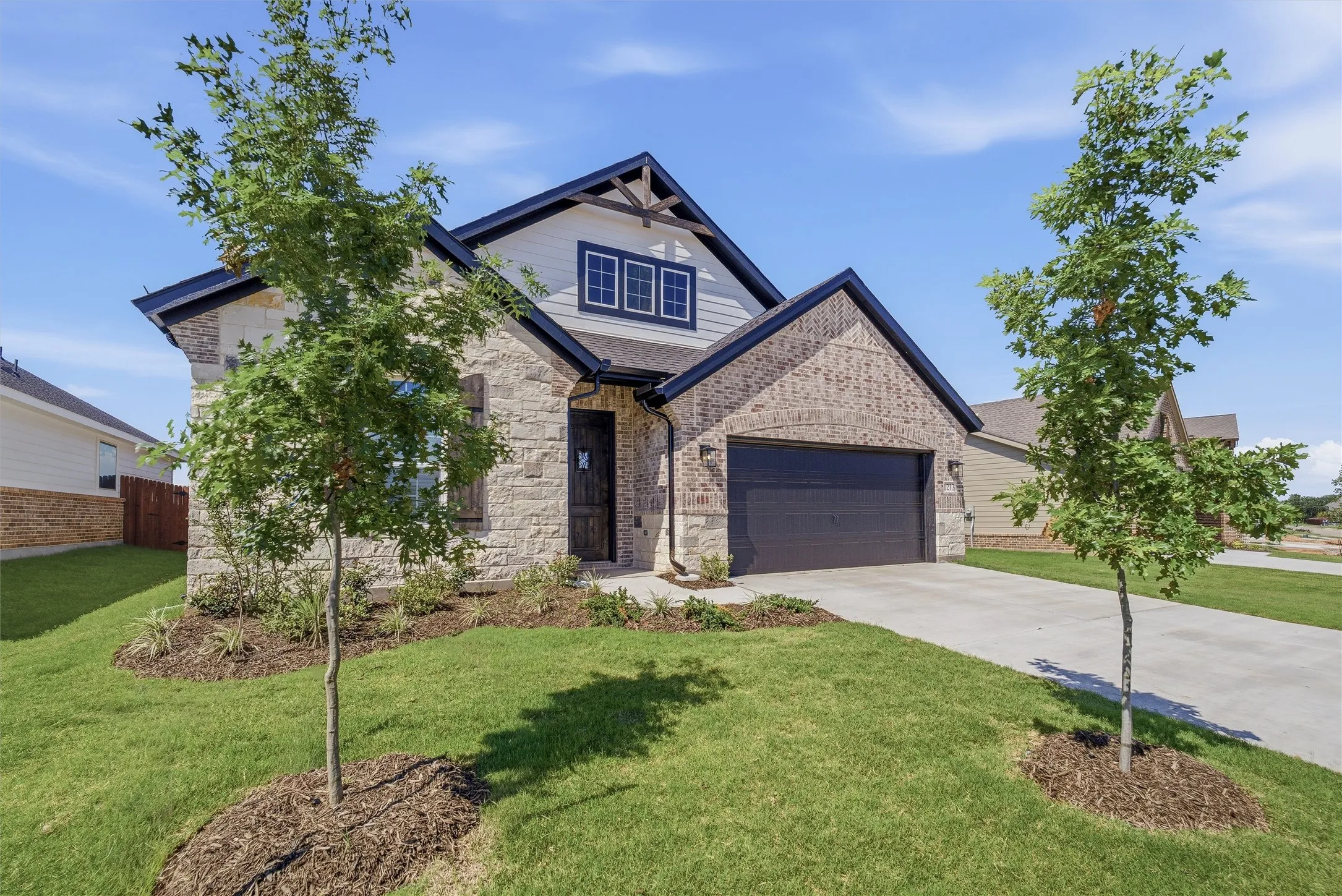 Stone and brick exterior with dark trim accents, featuring a two-car garage, established front landscaping, and a concrete driveway