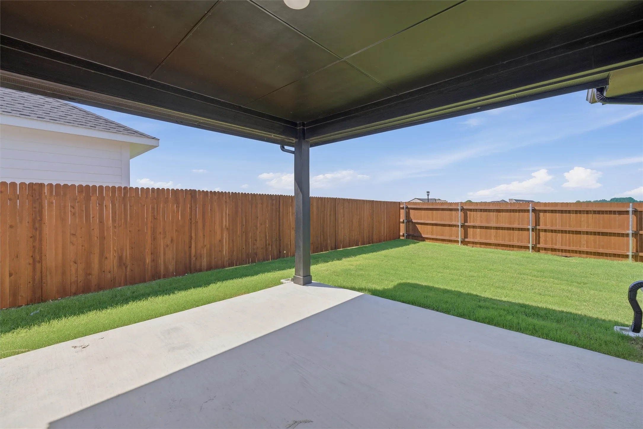 Covered patio with concrete flooring, overlooking a grass lawn enclosed by a wood privacy fence