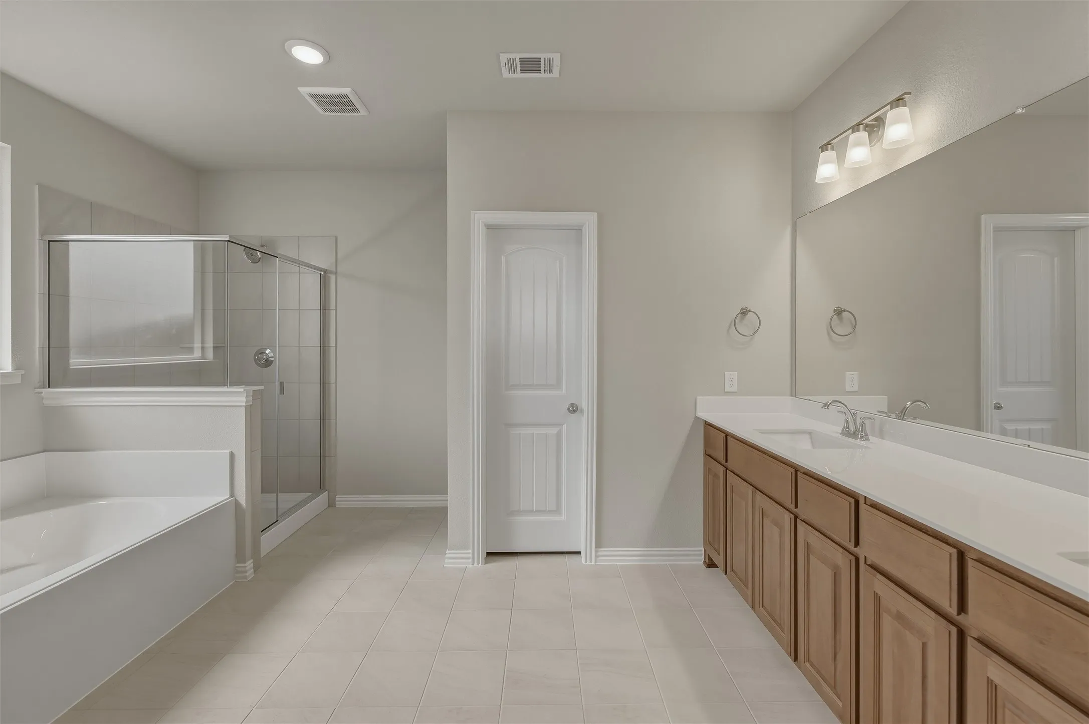 Spacious bathroom featuring a dual vanity with light-toned countertops, a glass-enclosed shower, a separate soaking tub, and neutral-toned floor tiling