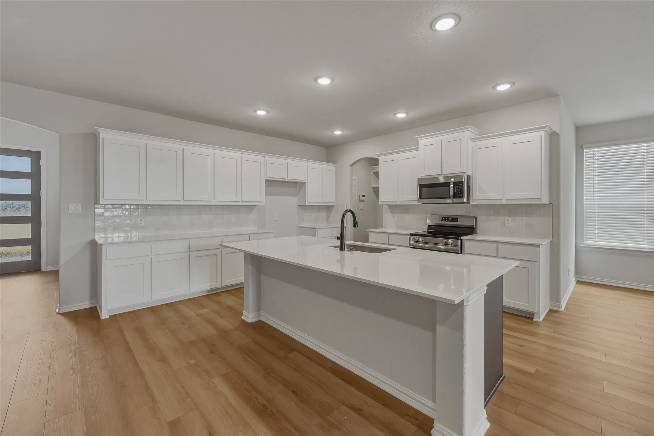 Kitchen featuring an expansive island with a built-in sink and dark-finish faucet, extensive white cabinetry, light-toned wood-finish flooring, recessed lighting, and stainless steel appliances