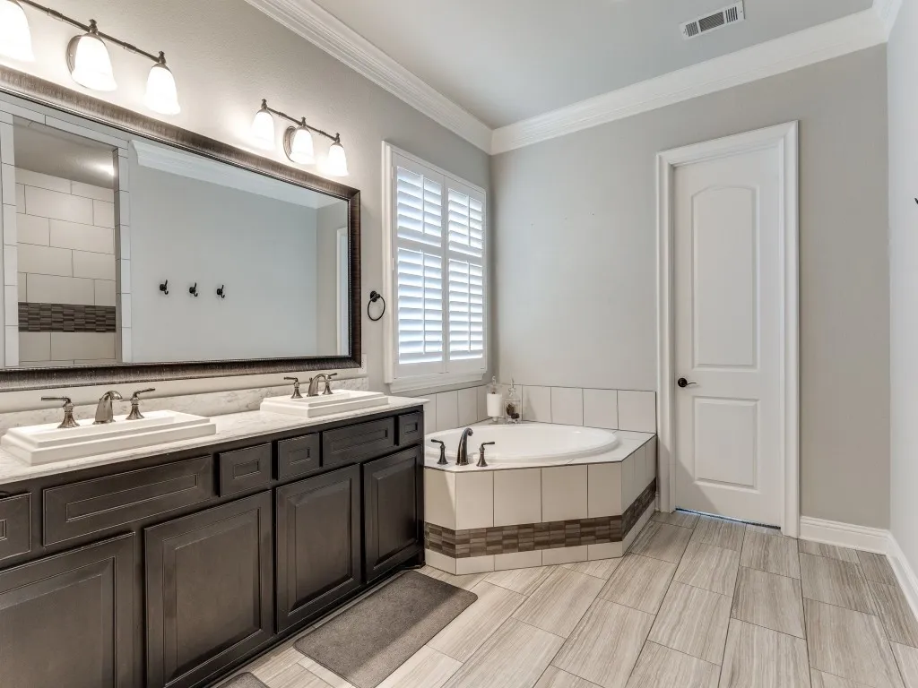 Bathroom featuring a dual vanity with rectangular sinks, a garden tub with tiled surround, wood-finish tile flooring, and plantation shutters