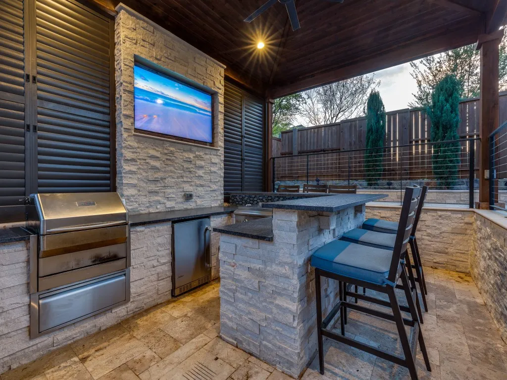 Outdoor kitchen featuring an integrated grill, under-counter refrigerator, stone-clad island with countertop, and stone accent wall with built-in television