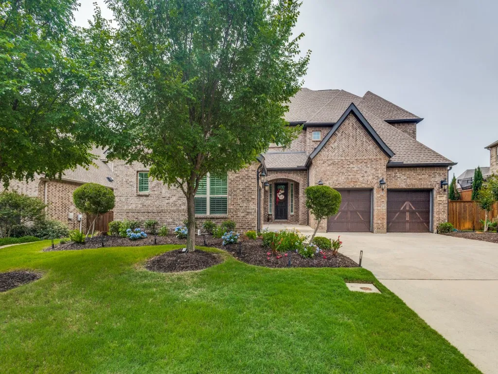 Brick facade residence with dark-paneled garage doors, a concrete driveway, and a landscaped front yard featuring mature trees and flower beds