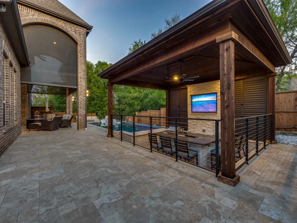Outdoor living space featuring a wood-beam pergola with a ceiling fan, stone-faced outdoor kitchen with integrated television, and sleek cable railing