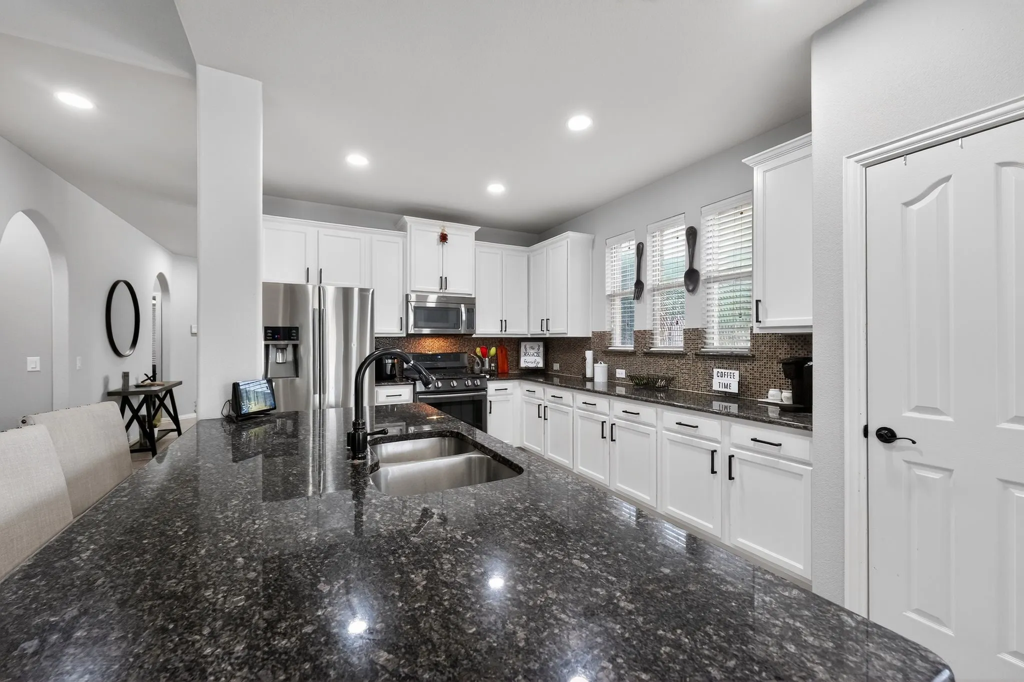 Spacious kitchen featuring a dark granite island, stainless steel appliances, white cabinetry, a large basin sink with a matte black faucet, and a mosaic tile backsplash