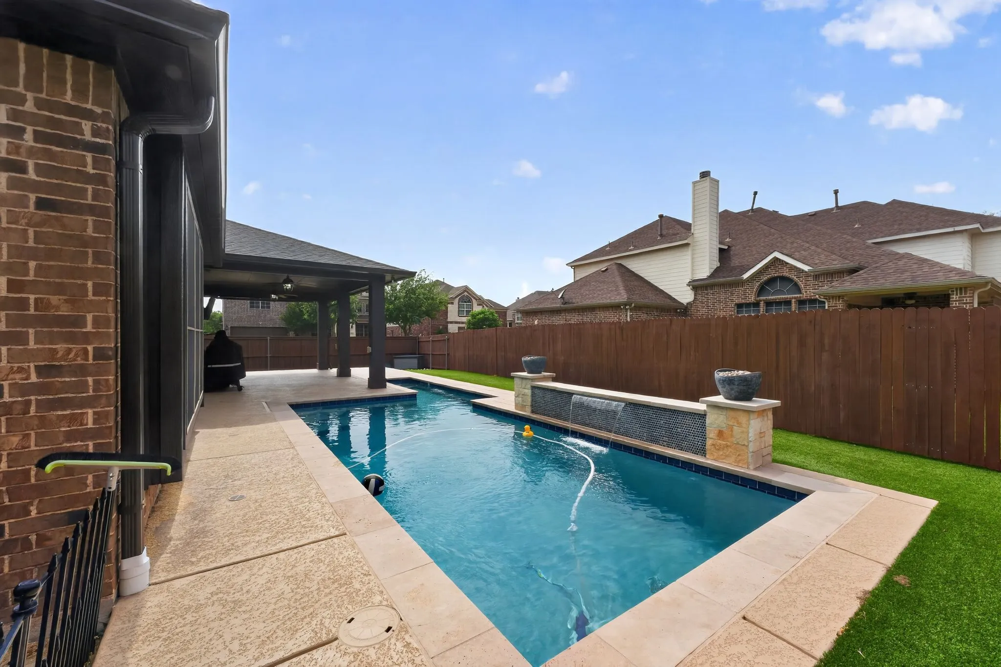 Backyard featuring a rectangular swimming pool with a water feature, a covered patio, and artificial turf