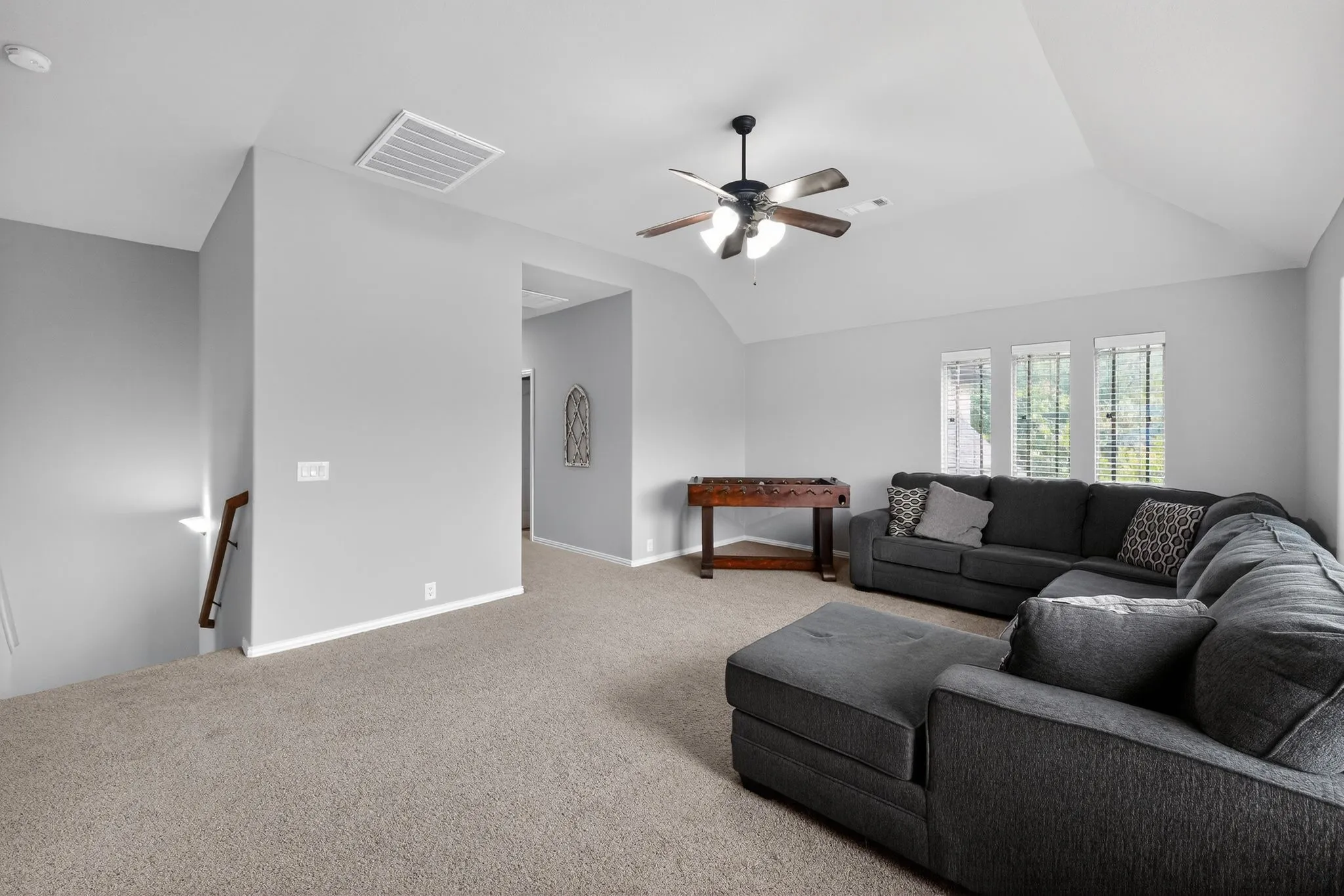 Upstairs spacious entertainment room featuring carpet, vaulted ceilings, a ceiling fan with light fixture, and triple windows with blinds