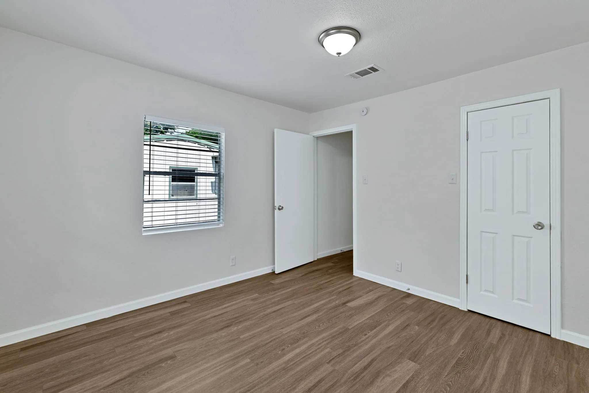 Expansive room featuring wood-finish flooring, light gray walls, a window with horizontal blinds, a ceiling-mounted light fixture, and two white paneled doors