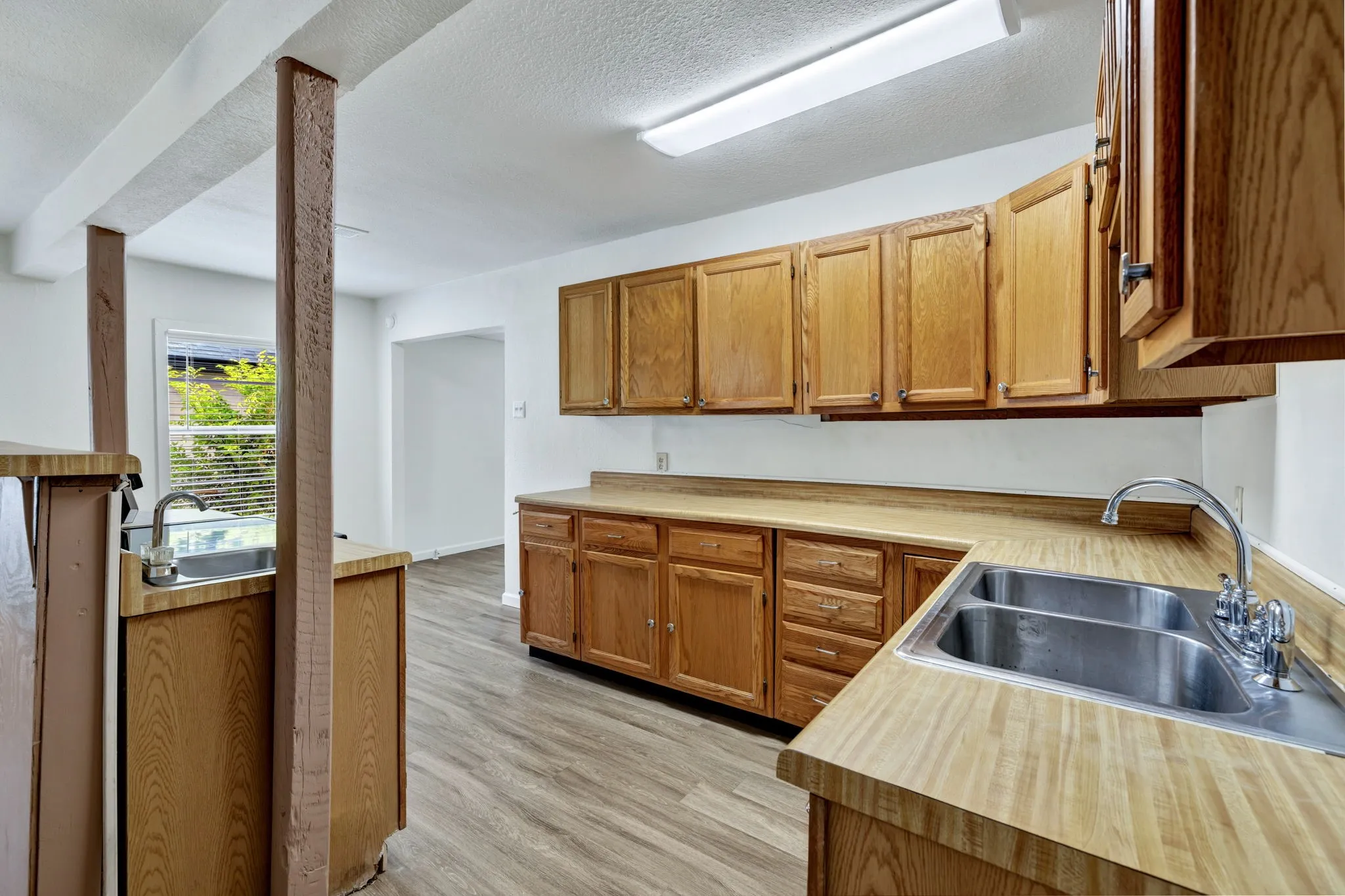 Kitchen area featuring wood-finish laminate flooring, light wood cabinetry, laminate countertops, a double basin stainless steel sink with a chrome faucet, and exposed support beams