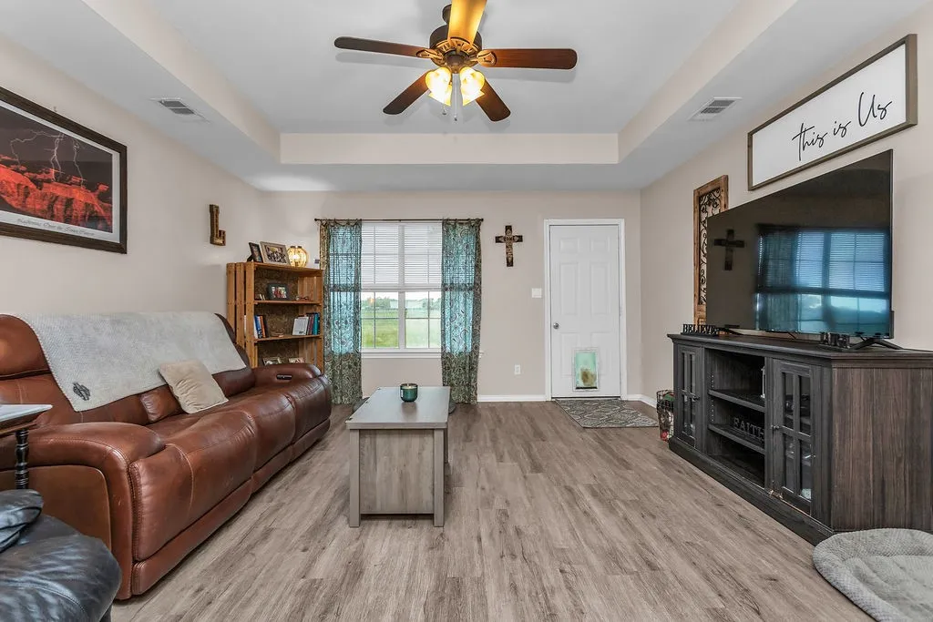 Living area featuring wood-finish flooring, a tray ceiling with a ceiling fan, and a double window with blinds