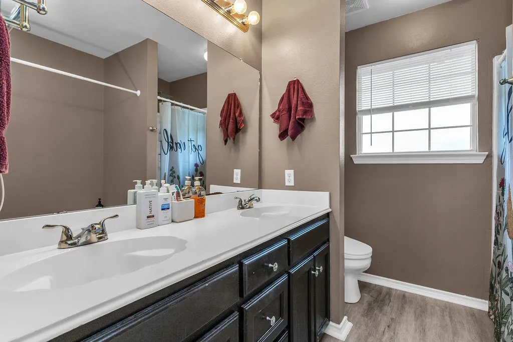 Bathroom featuring a dual vanity with dark wood cabinetry and white countertops, a large mirror, a window with horizontal blinds, and wood-finish flooring
