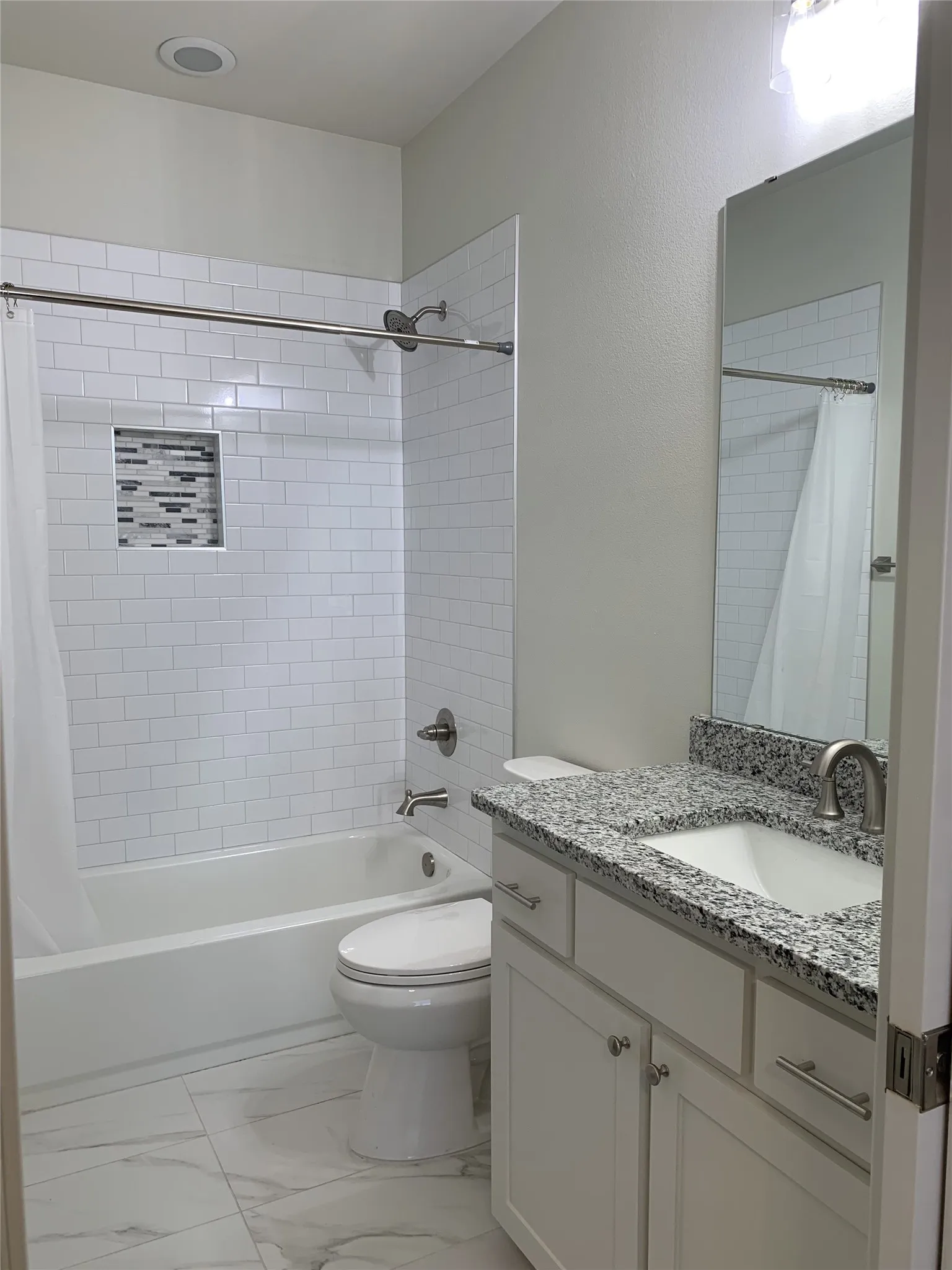 Bathroom featuring a white subway tile tub surround with an accent niche, a white vanity with a speckled countertop, an undermount sink, and marble-look floor tiling